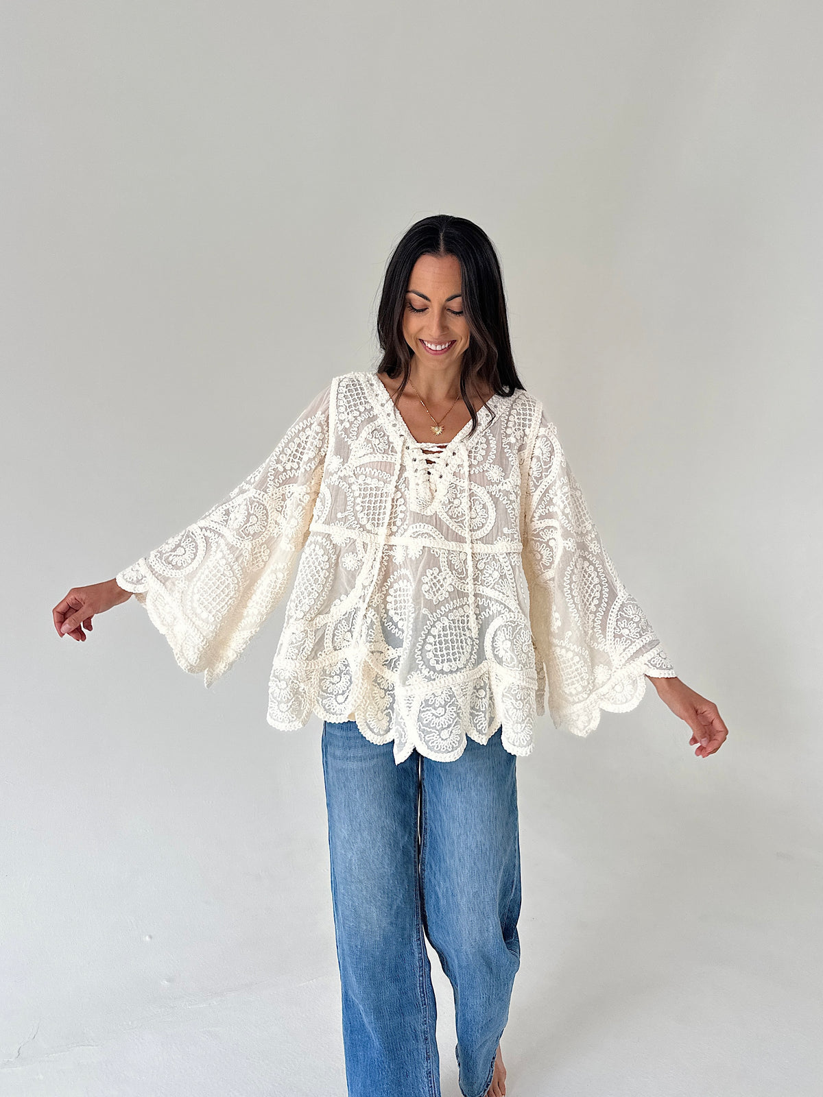 A woman modeling a cream lace bohemian-style top with wide bell sleeves and blue jeans, standing against a plain light gray studio background.