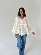 A woman wearing a cream lace bohemian-style long-sleeve top and blue jeans, standing against a plain light gray studio background.