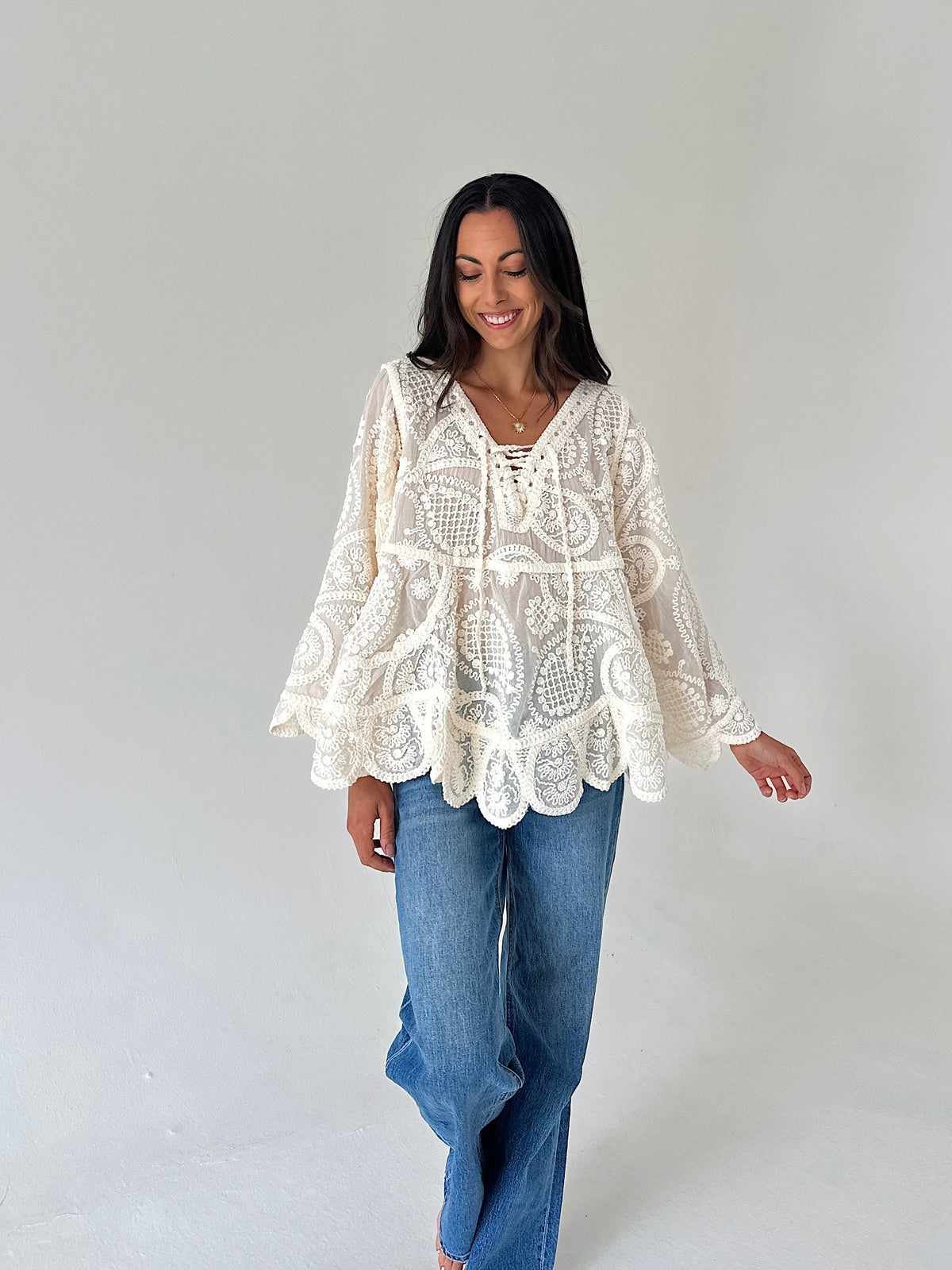 A woman wearing a cream lace bohemian-style long-sleeve top and blue jeans, standing against a plain light gray studio background.