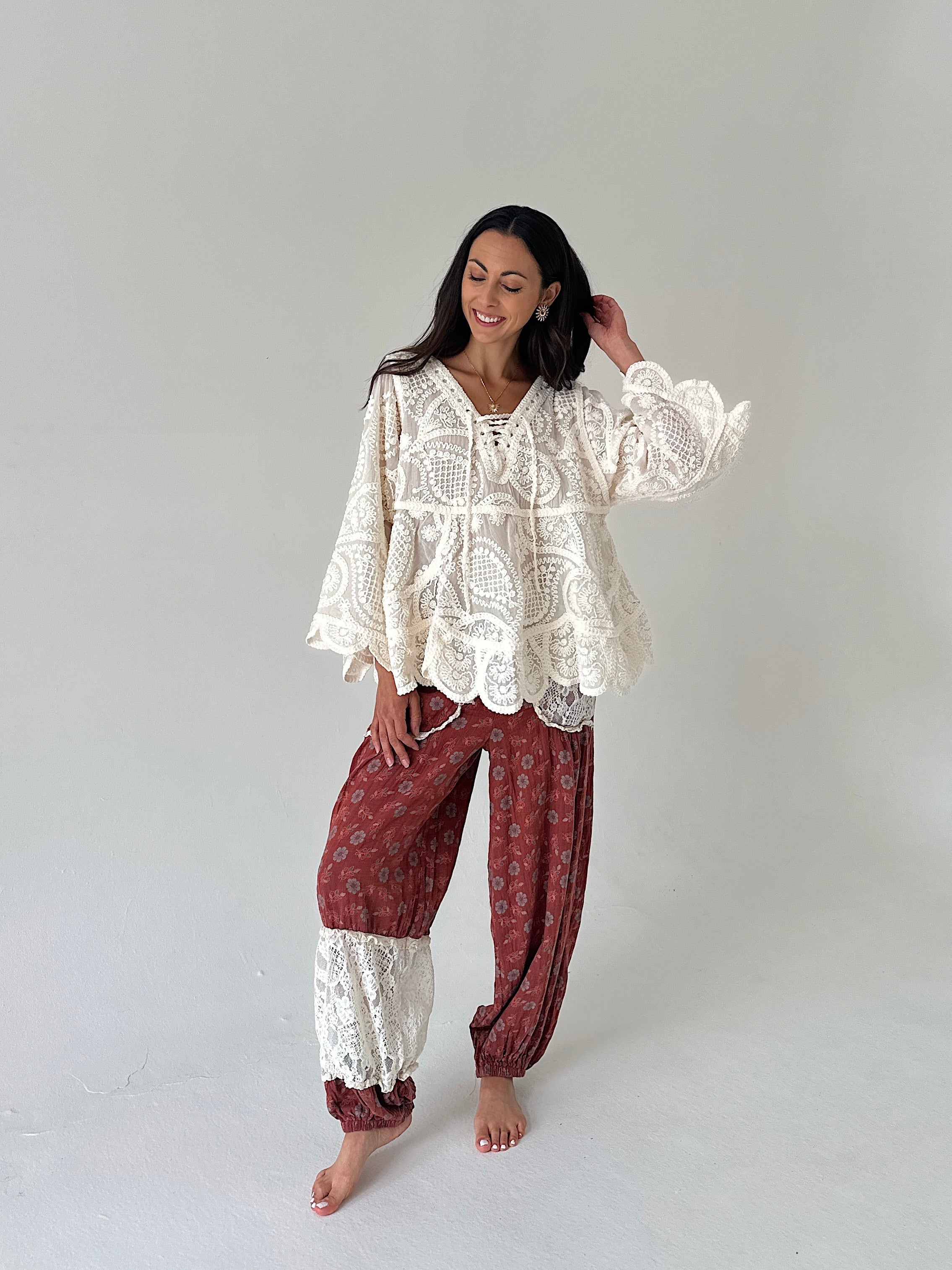 A woman modeling a bohemian-style two-piece outfit consisting of a cream lace long-sleeve top and rust-red patterned harem pants with lace cuffs, standing against a plain light gray studio background.