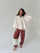 A woman modeling a bohemian-style two-piece outfit consisting of a cream lace long-sleeve top and rust-red patterned harem pants with lace cuffs, standing against a plain light gray studio background.