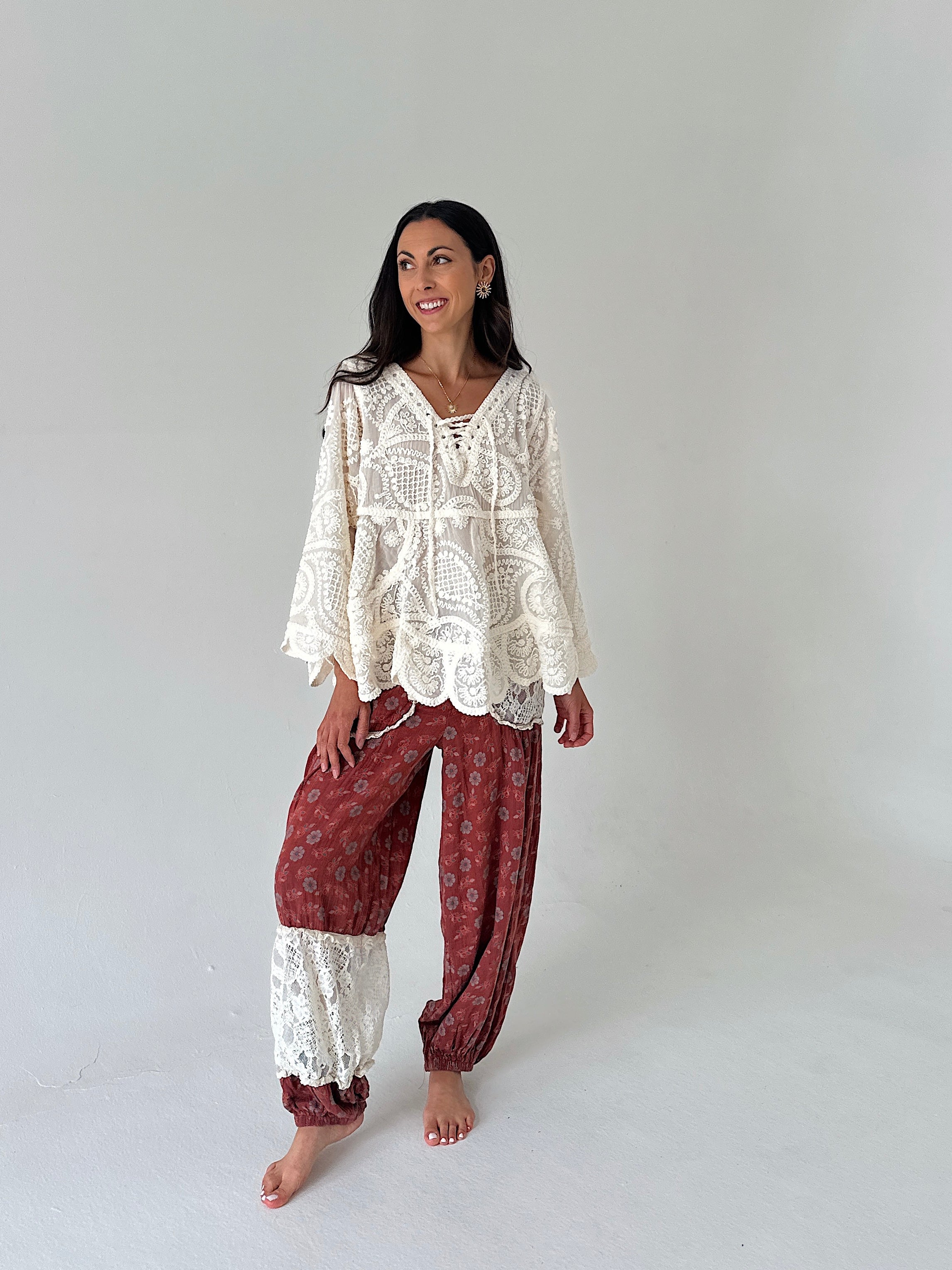 A woman modeling a bohemian-style two-piece outfit consisting of a cream lace long-sleeve top and rust-red patterned harem pants with lace cuffs, standing against a plain light gray studio background.