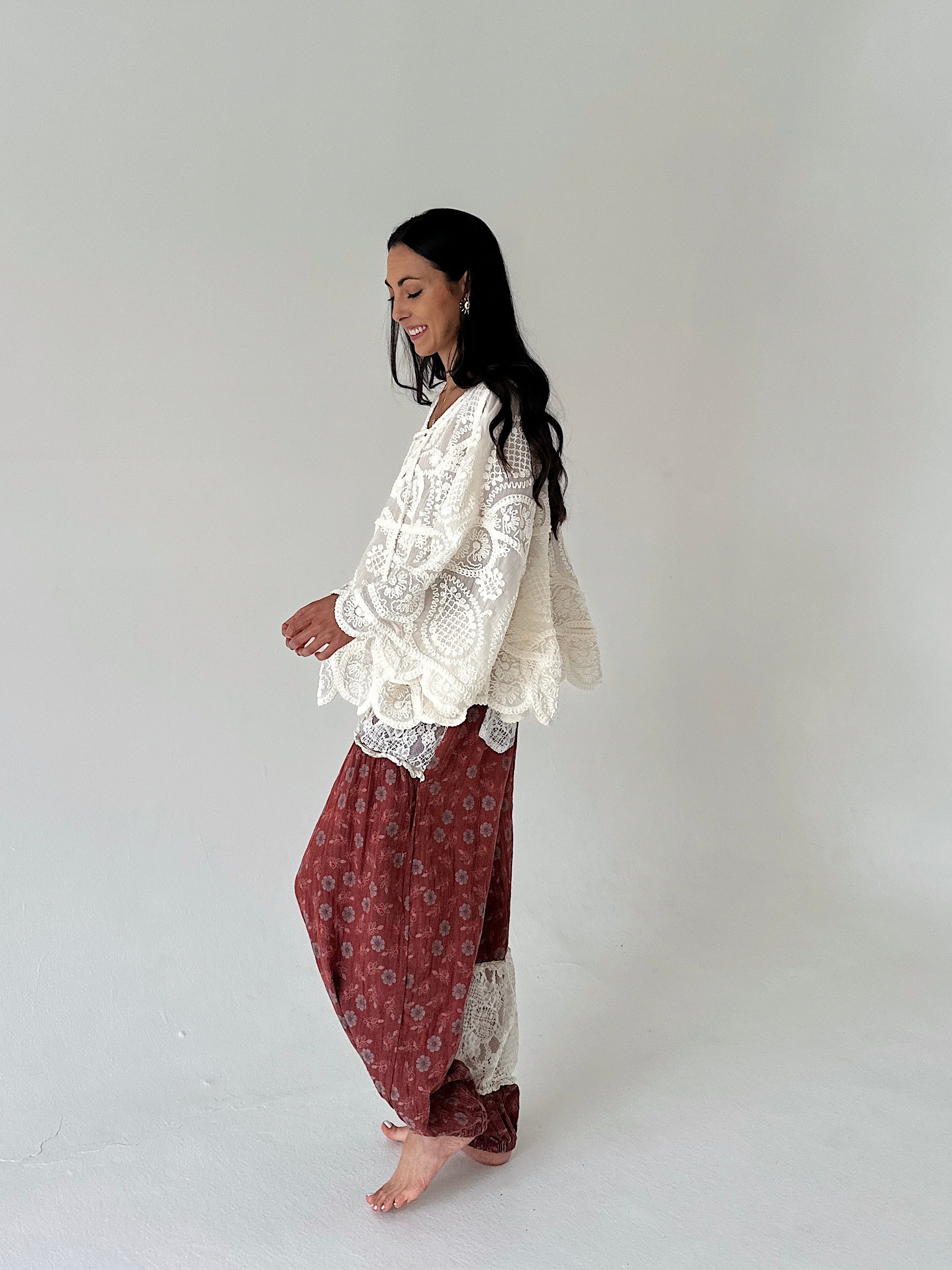 A woman stands barefoot in profile against a plain light gray studio background, wearing a loose white lace top and rust-red patterned harem-style pants.