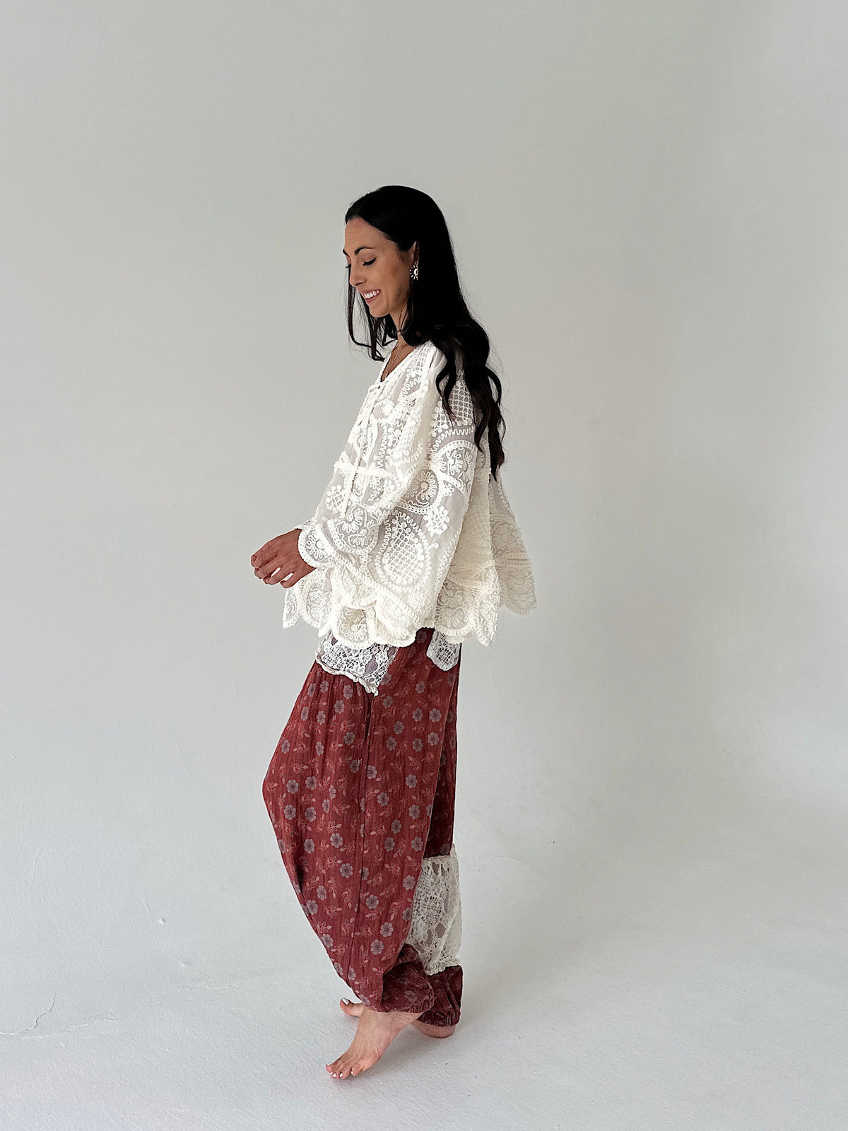 A woman stands barefoot in profile against a plain light gray studio background, wearing a loose white lace top and rust-red patterned harem-style pants.