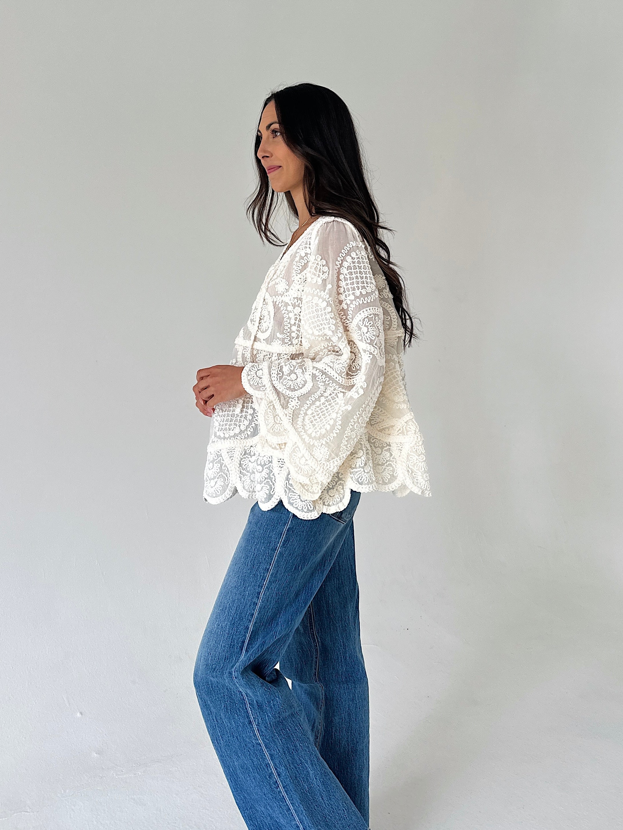 A woman wearing a cream-colored, long-sleeved lace blouse and wide-leg blue jeans, standing in profile against a plain light gray studio background.