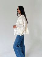 A woman wearing a cream-colored, long-sleeved lace blouse and wide-leg blue jeans, standing in profile against a plain light gray studio background.