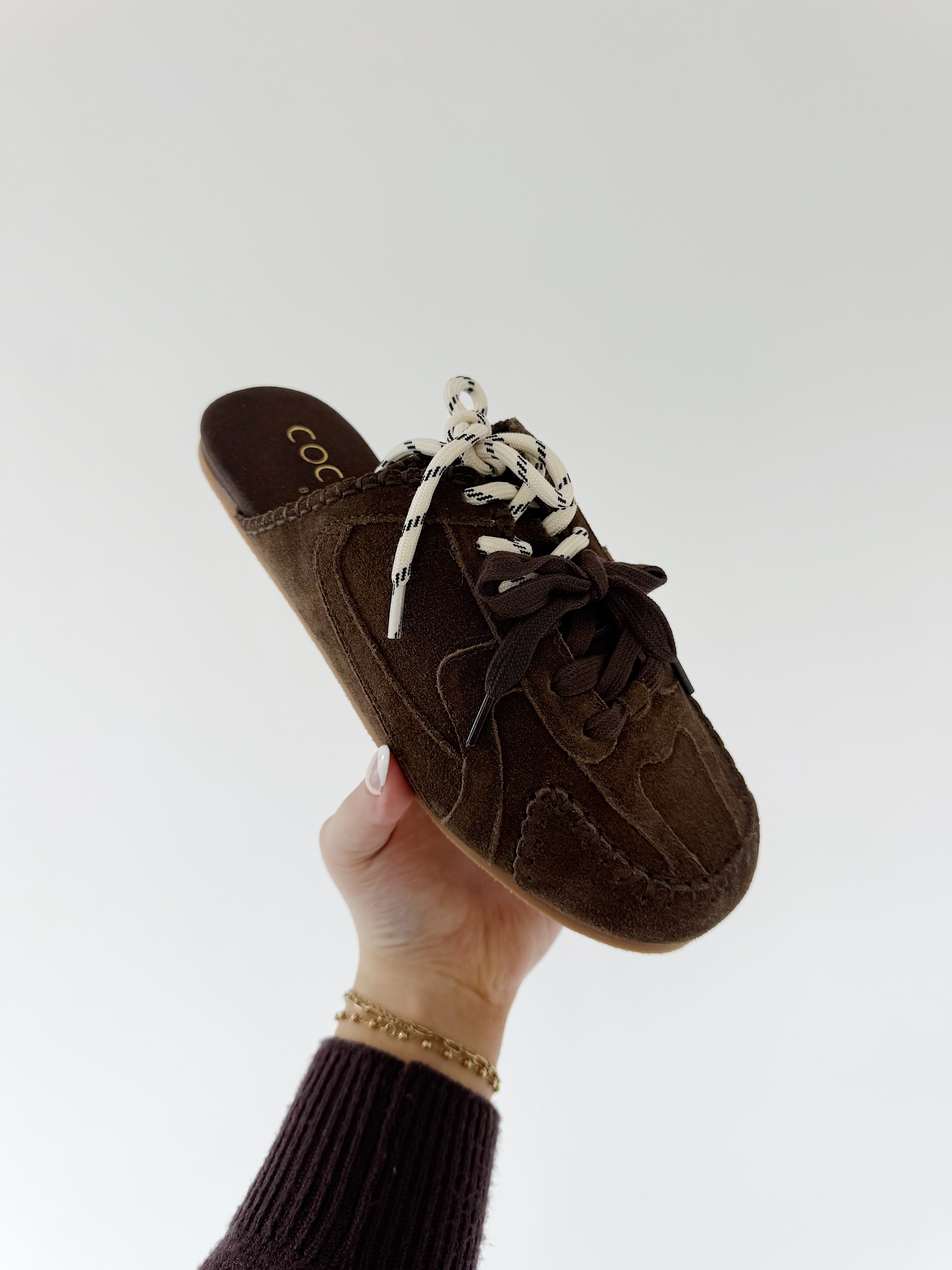 Dark brown sneaker mule shoe with white laces held by a hand against a white background