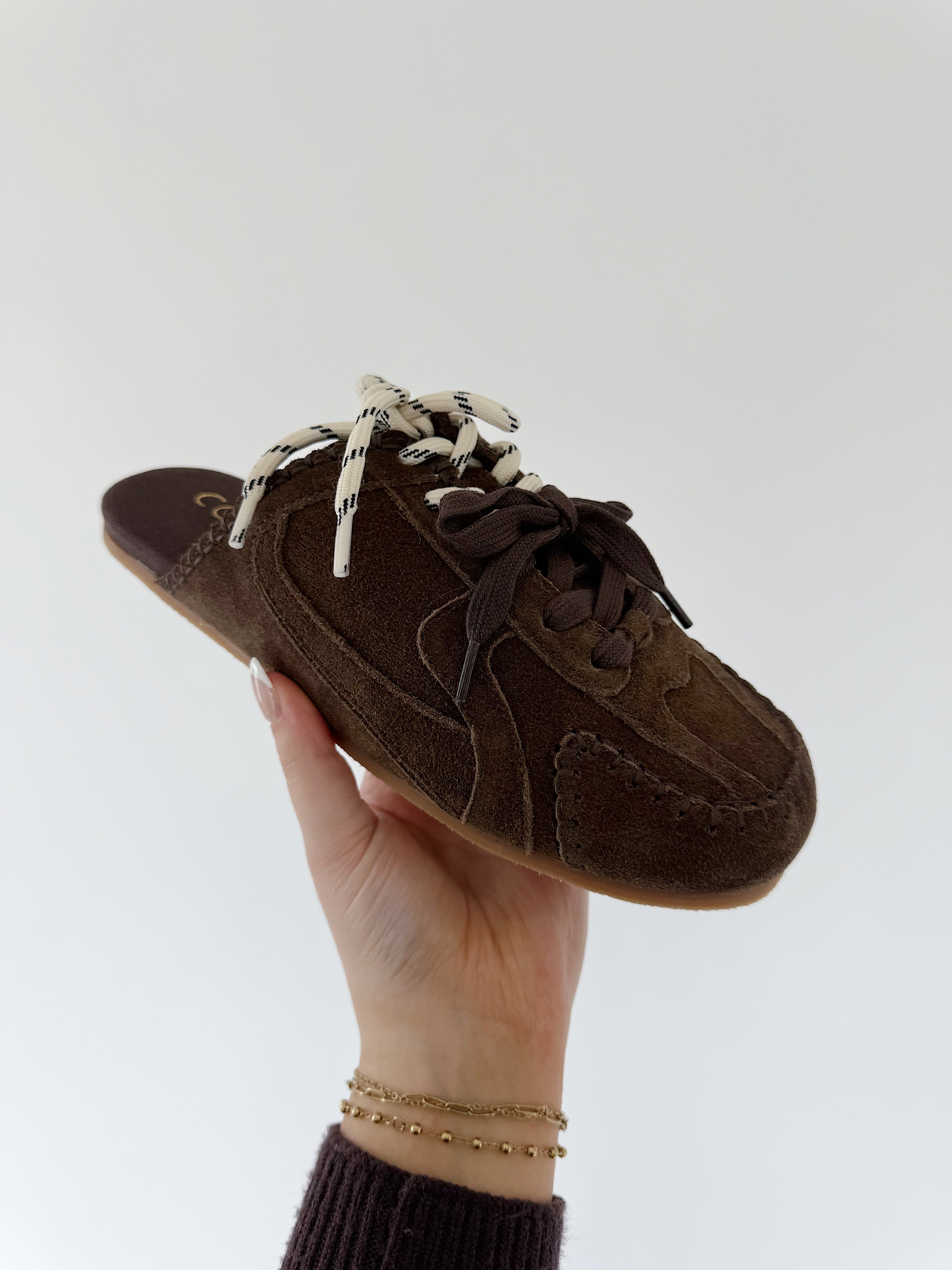 Brown suede sneaker slide shoe with white laces held by a hand against a light gray background
