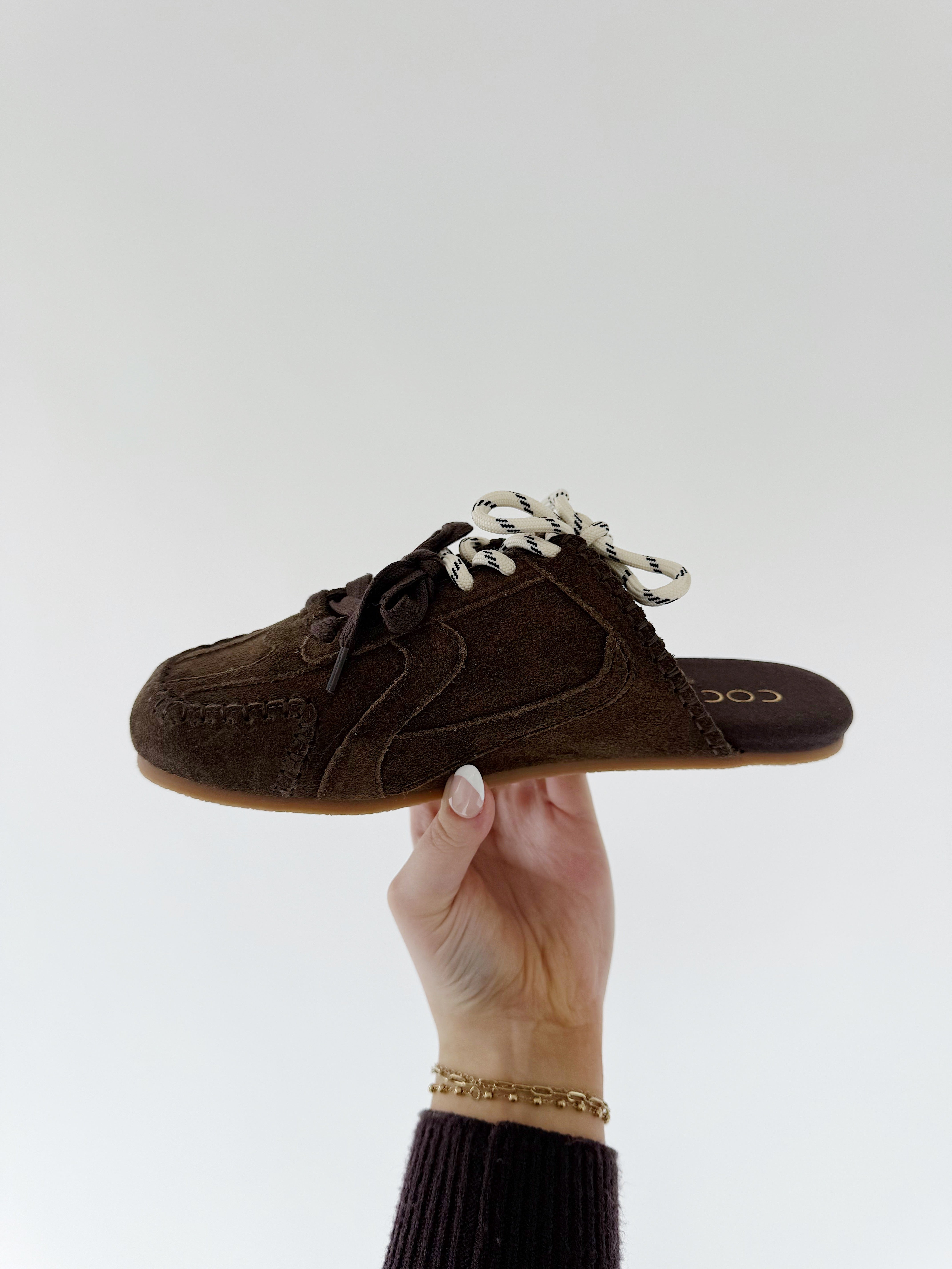 Brown suede sneaker mule shoe held by a hand against a white background