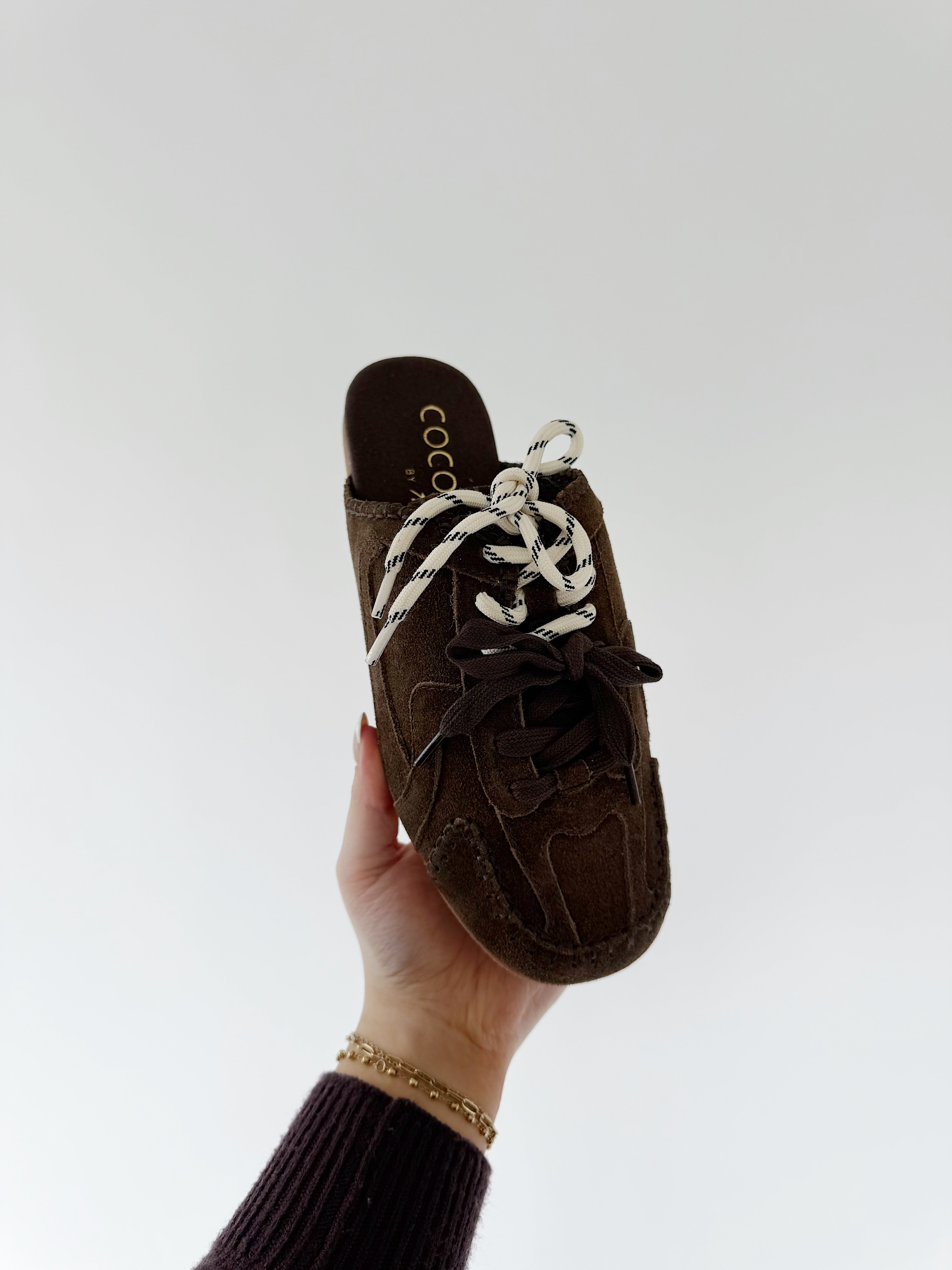 Brown slip-on shoe with white laces held by a hand on a light gray background
