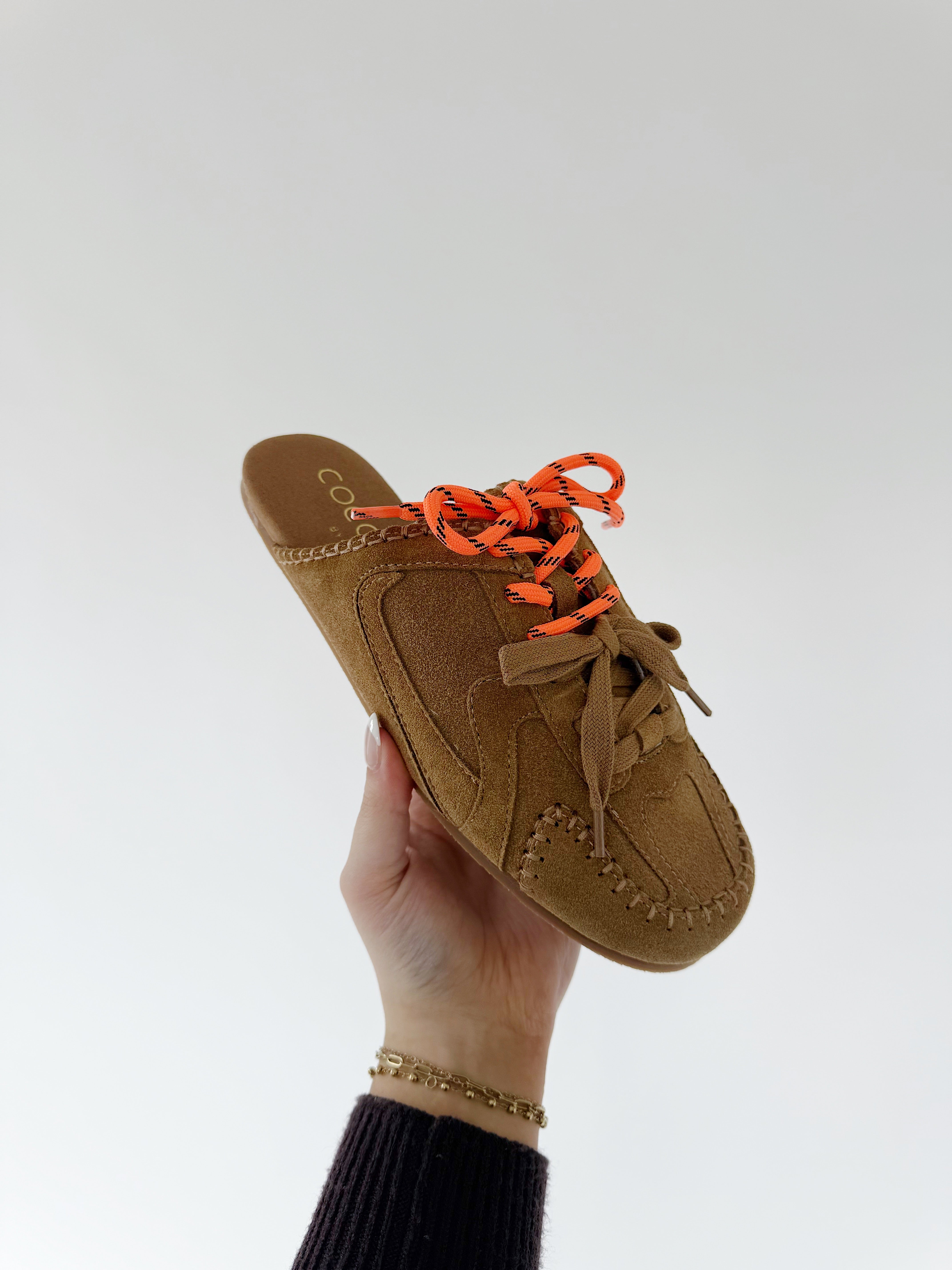 Brown slip-on shoe with orange laces held by a hand against a white background