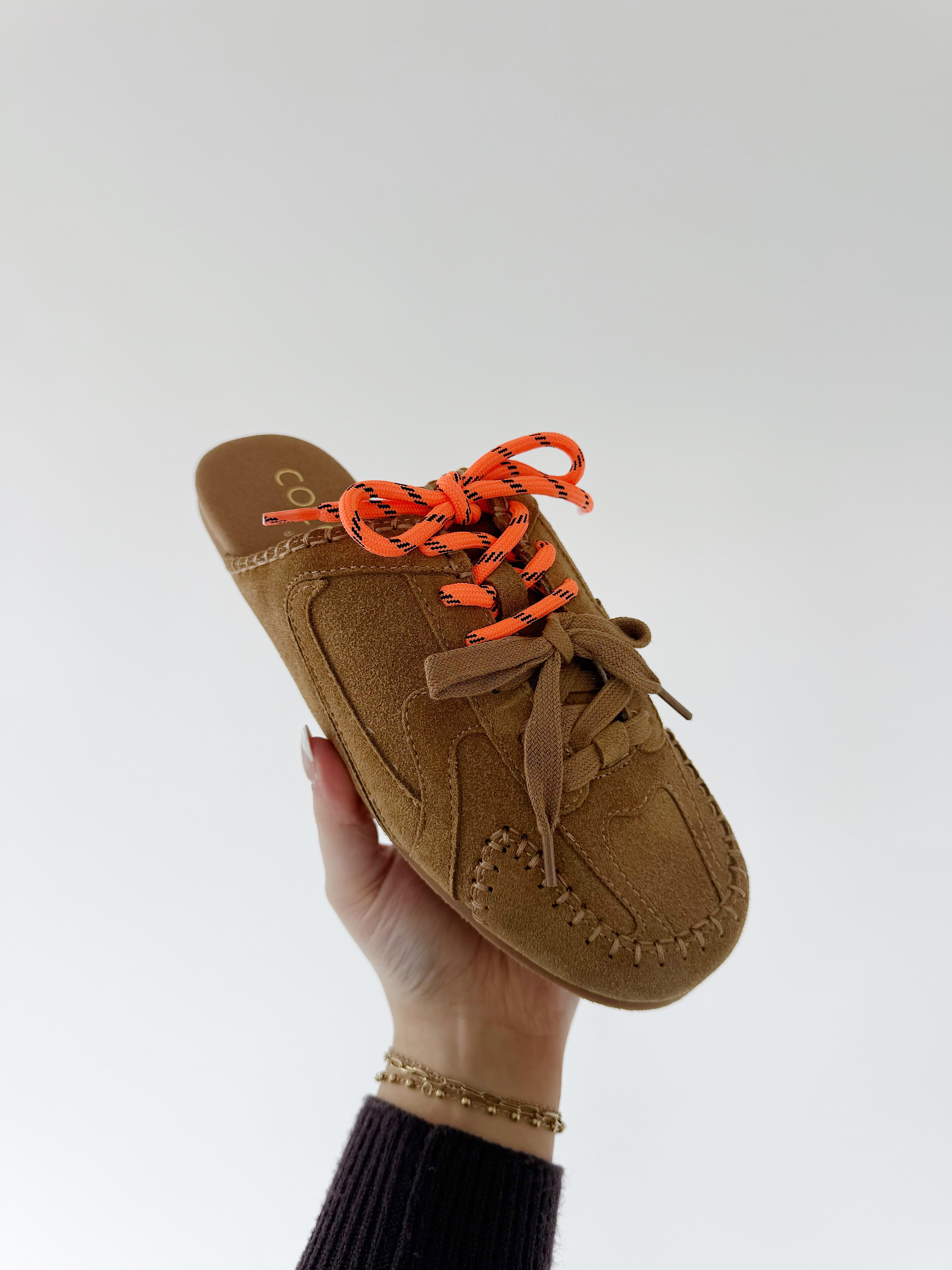 Brown suede shoe with orange laces held by a hand against a white background