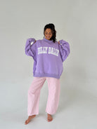 Woman wearing a purple 'Dilly Dally' sweatshirt and pink checkered pants on a white background