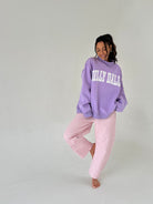 Woman wearing a purple 'Dilly Dally' sweatshirt and pink checkered pants on a plain background