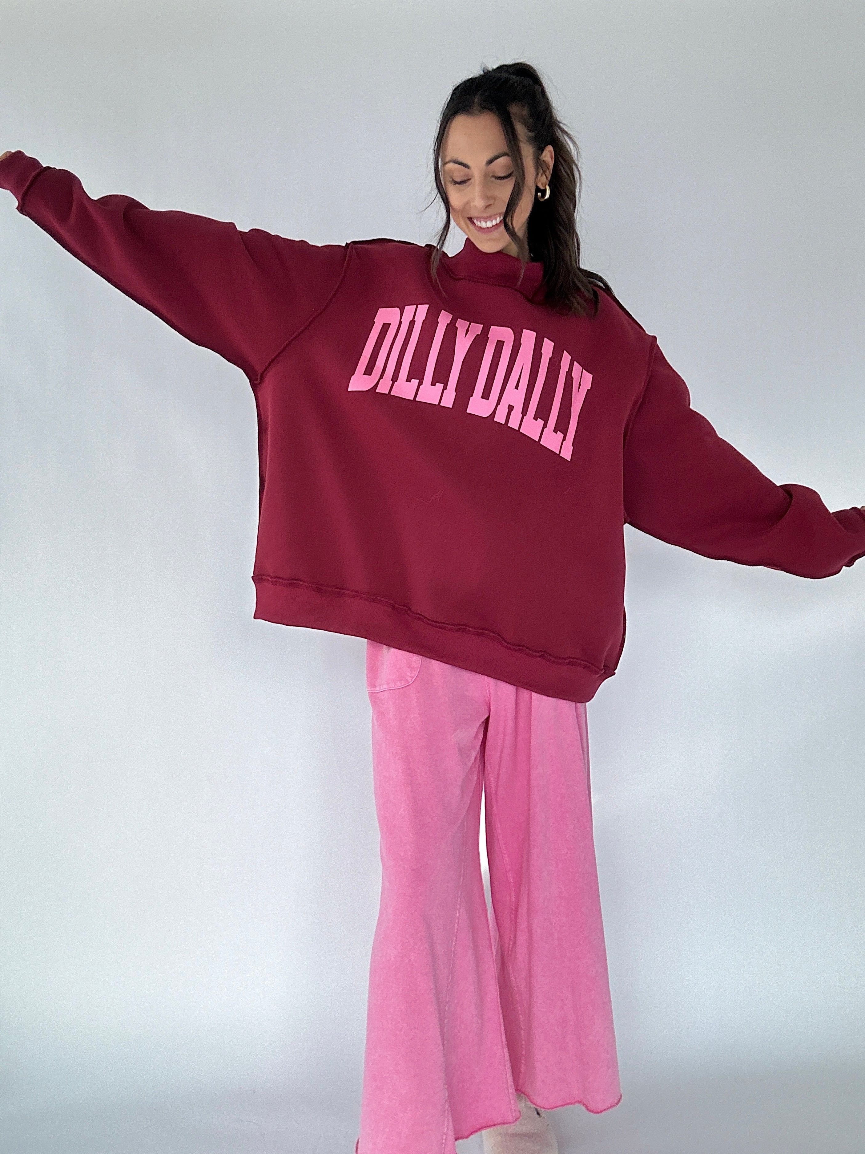 Person wearing a maroon mockneck pullover with pink 'DILLY DALLY' text and pink pants on a white background
