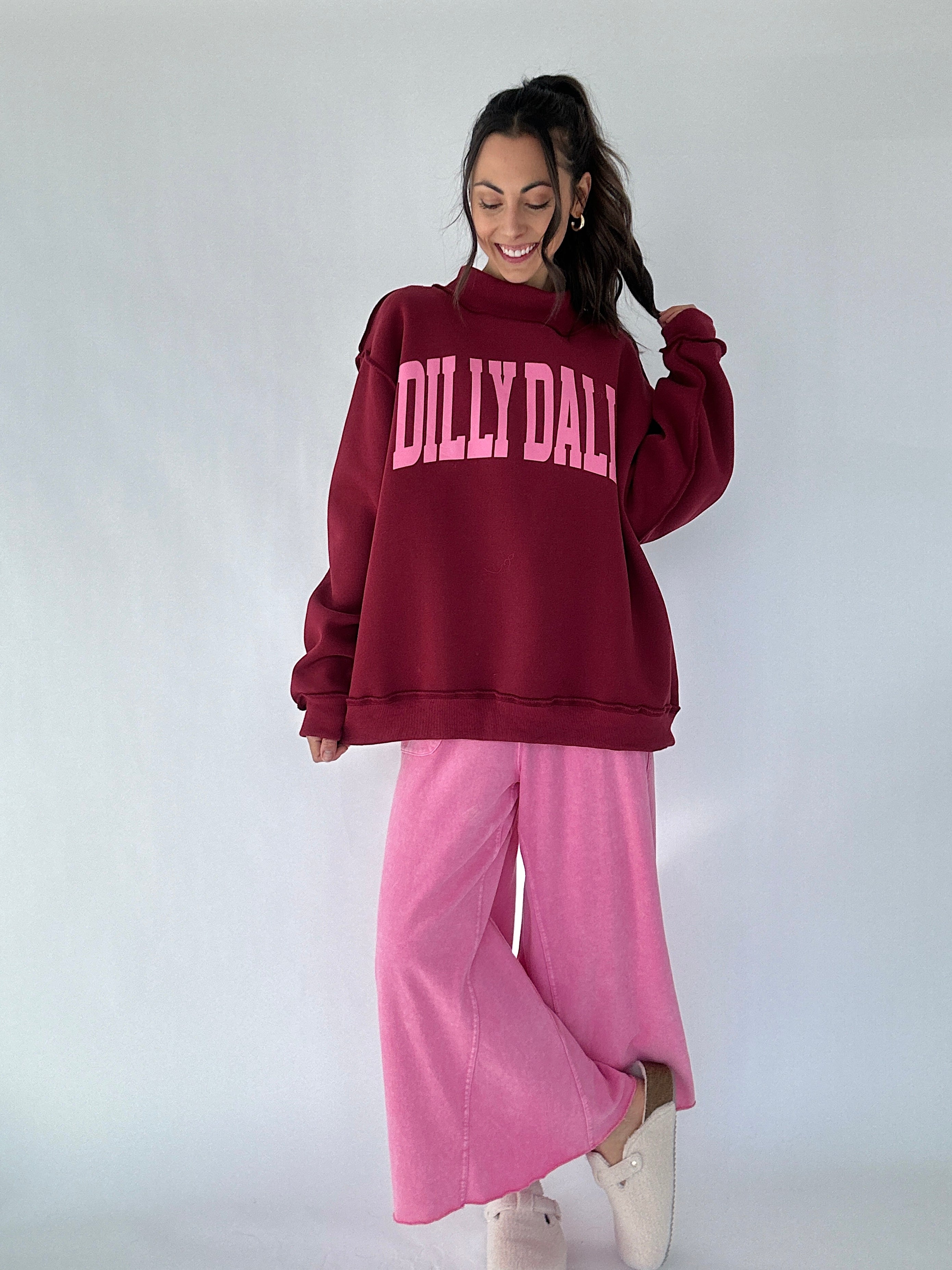 Person wearing a maroon 'Dilly Dally' mock neck pullover and pink pants on a white background