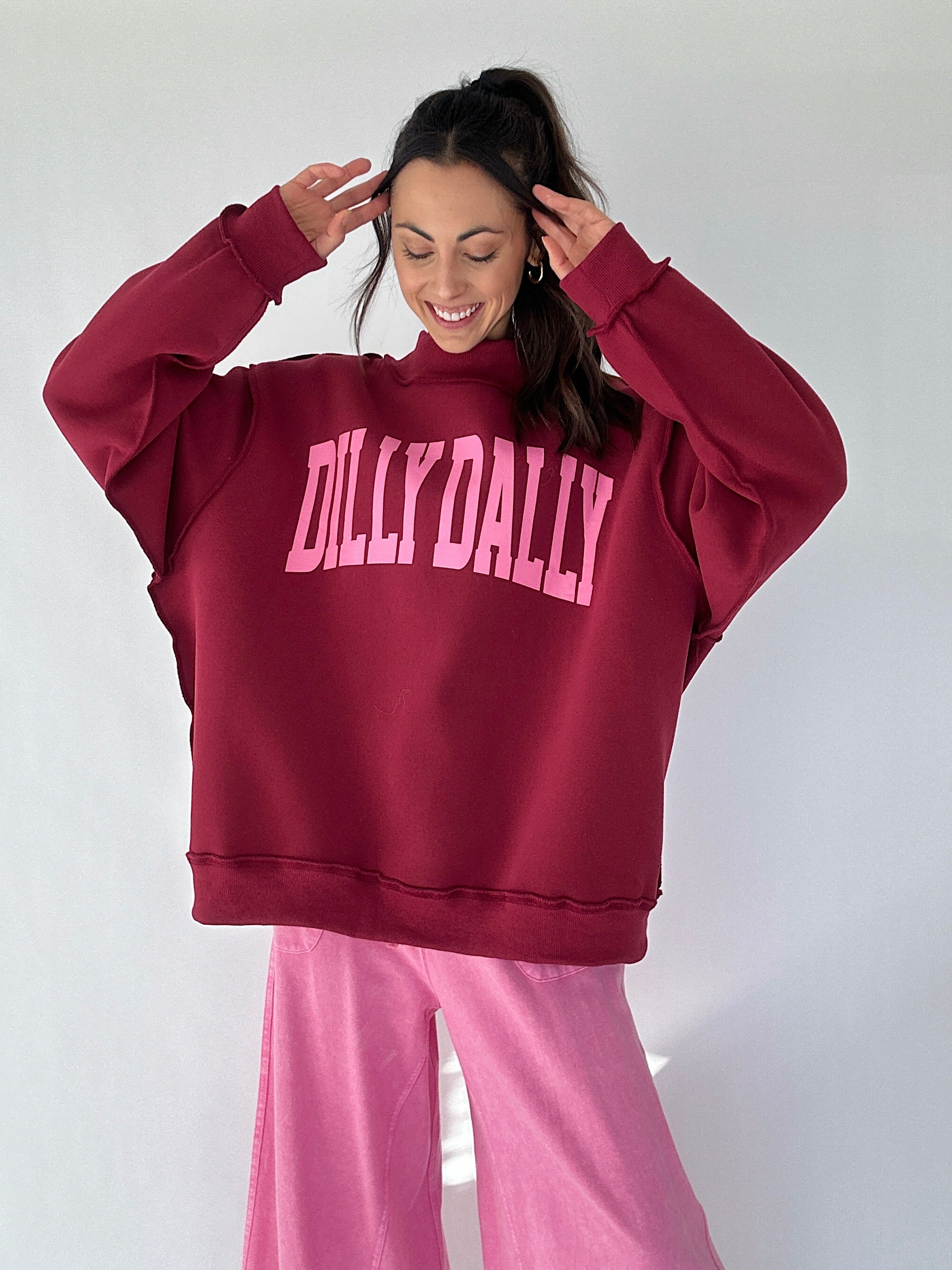 Person wearing a maroon mock neck pullover with 'DILLY DALLY' text and pink pants on a white background