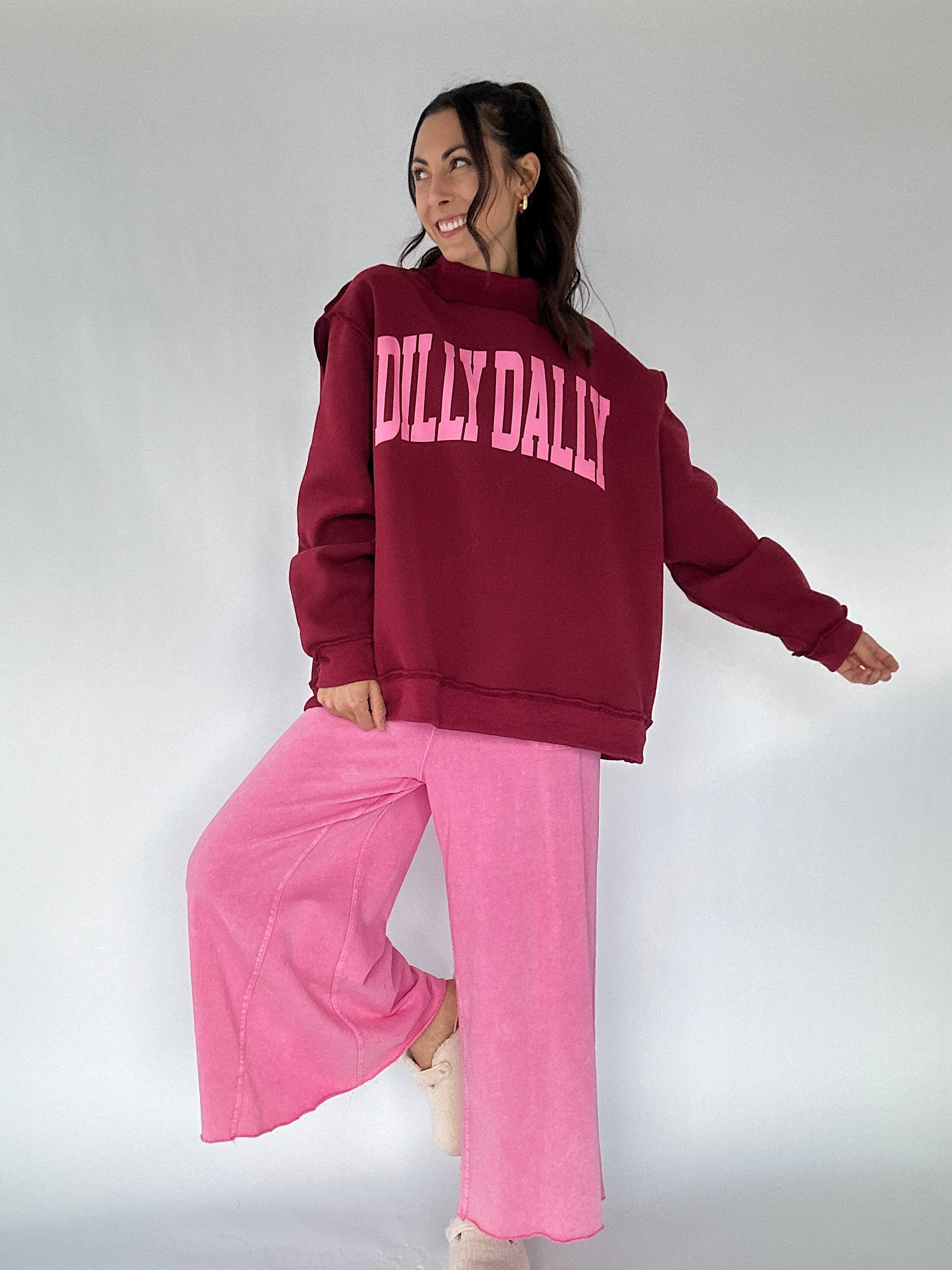 Person wearing a maroon 'DILLY DALLY' sweatshirt and pink pants on a white background