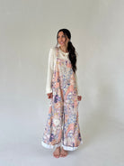 A woman stands barefoot against a plain light gray studio background, wearing a cream long-sleeve top layered under a wide-leg floral jumpsuit in pastel pink, purple, and beige tones.