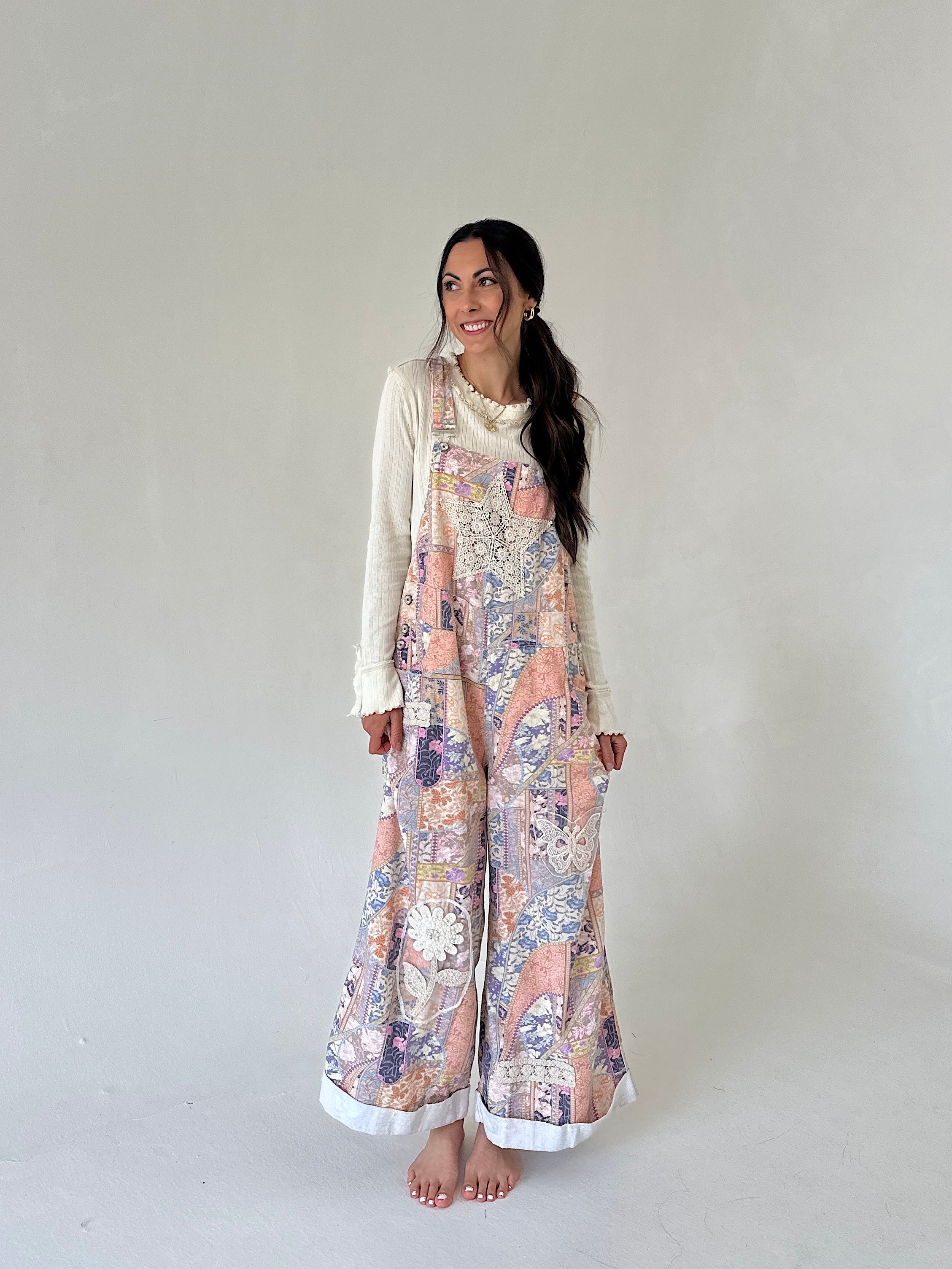A woman stands barefoot against a plain light gray studio background, wearing a cream long-sleeve top layered under a wide-leg floral jumpsuit in pastel pink, purple, and beige tones.