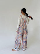 A woman stands barefoot against a plain off-white studio background, photographed from the back. She is wearing a long-sleeve cream top under loose, wide-leg overalls with a pastel multicolor patchwork print in pink, blue, beige, and purple tones.