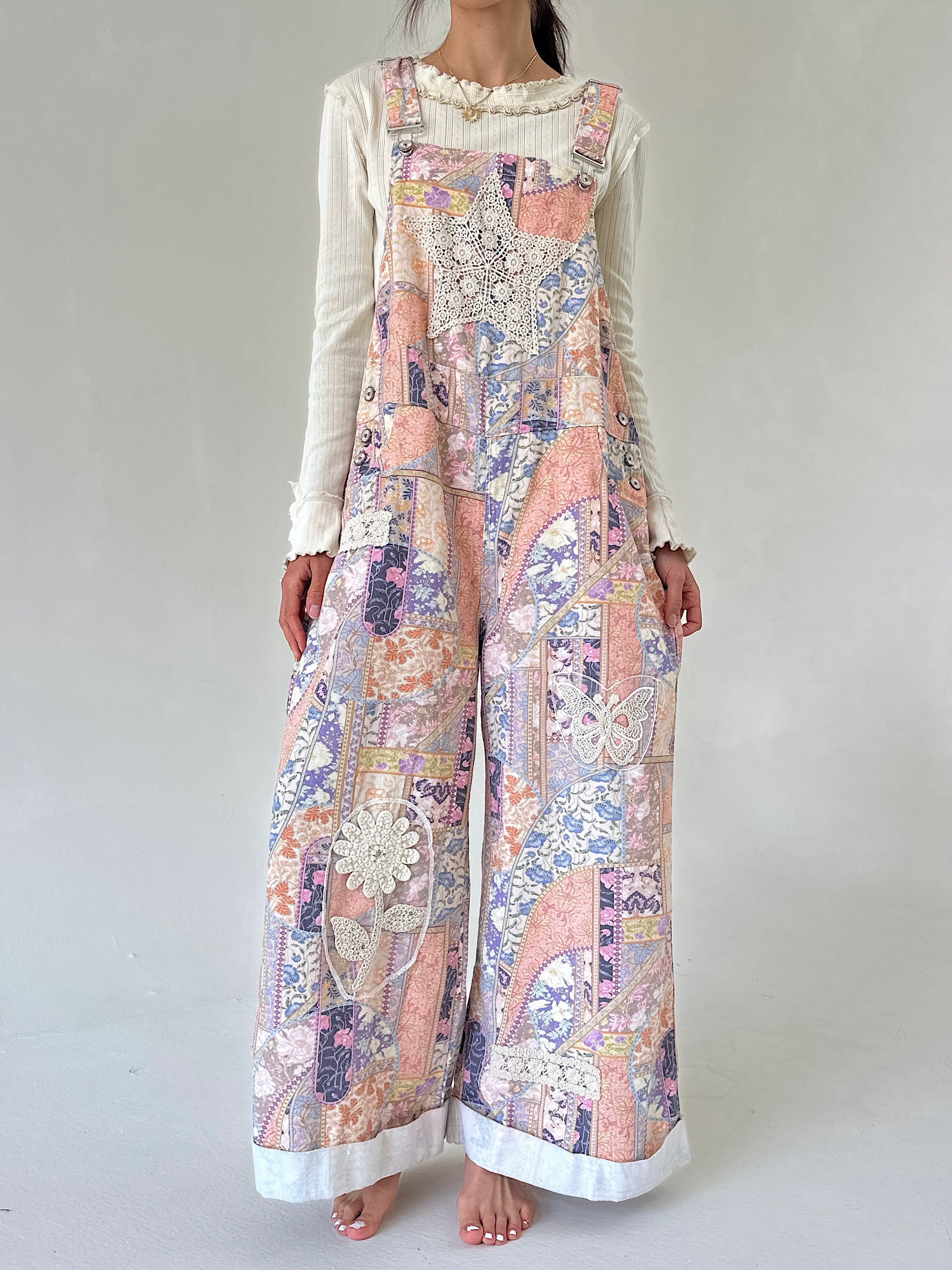 A woman modeling a lwide-leg overall jumpsuit with a multicolor patchwork print in pastel pink, purple, blue, and beige, layered over a white long-sleeve ruffled shirt, against a plain light gray background.