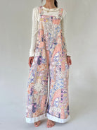 A woman modeling a lwide-leg overall jumpsuit with a multicolor patchwork print in pastel pink, purple, blue, and beige, layered over a white long-sleeve ruffled shirt, against a plain light gray background.