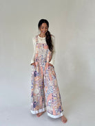 A woman modeling a sleeveless floral print overall jumpsuit with wide legs, worn over a cream long-sleeve top, standing against a plain light gray studio background.