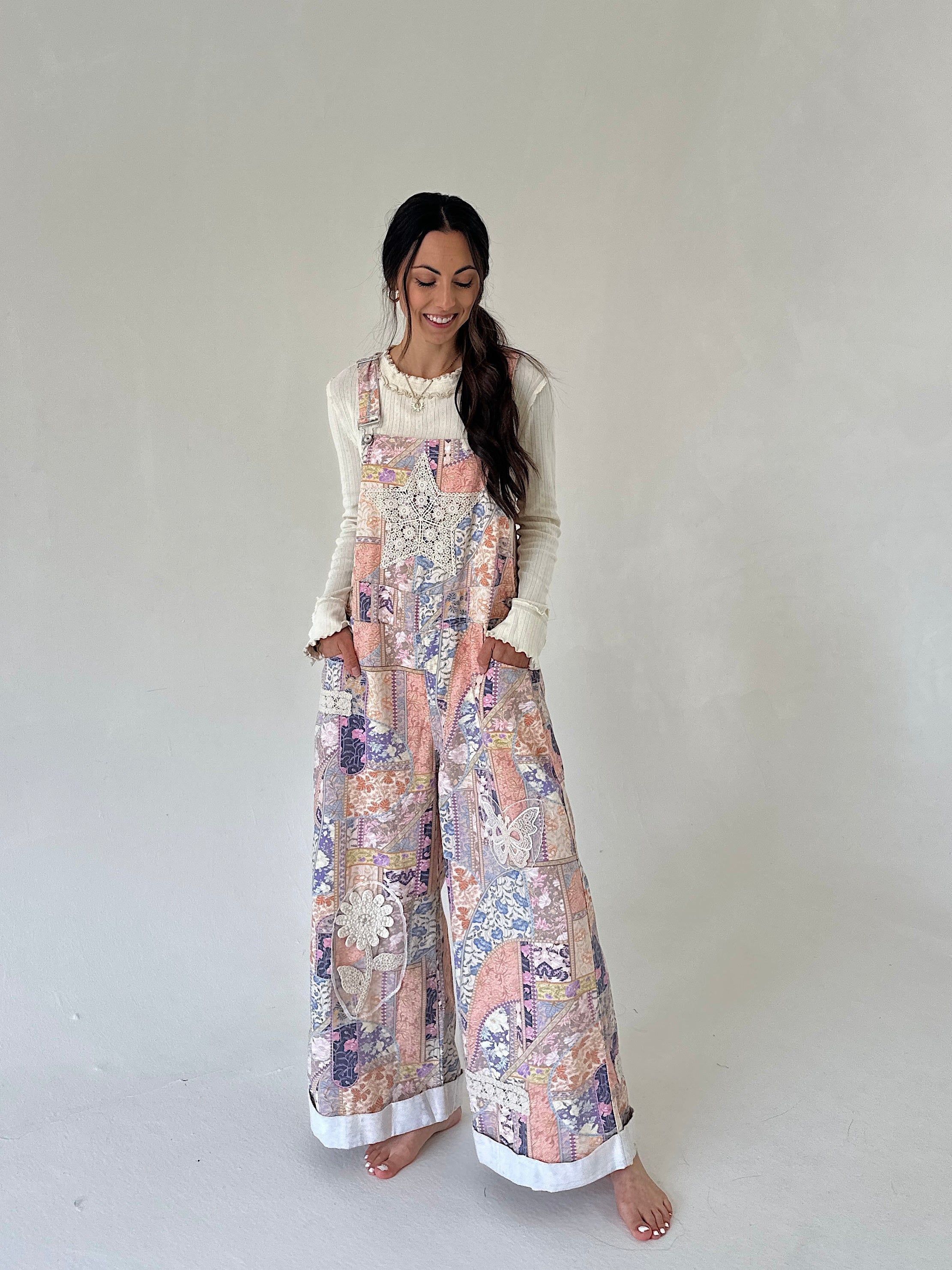 A woman modeling a sleeveless floral print overall jumpsuit with wide legs, worn over a cream long-sleeve top, standing against a plain light gray studio background.