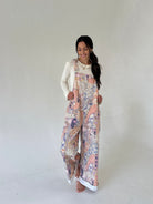 A woman modeling a pair of wide-leg jumpsuit overalls with a multicolor paisley and floral print in pink, beige, and purple, worn over a white long-sleeve top against a neutral studio background.