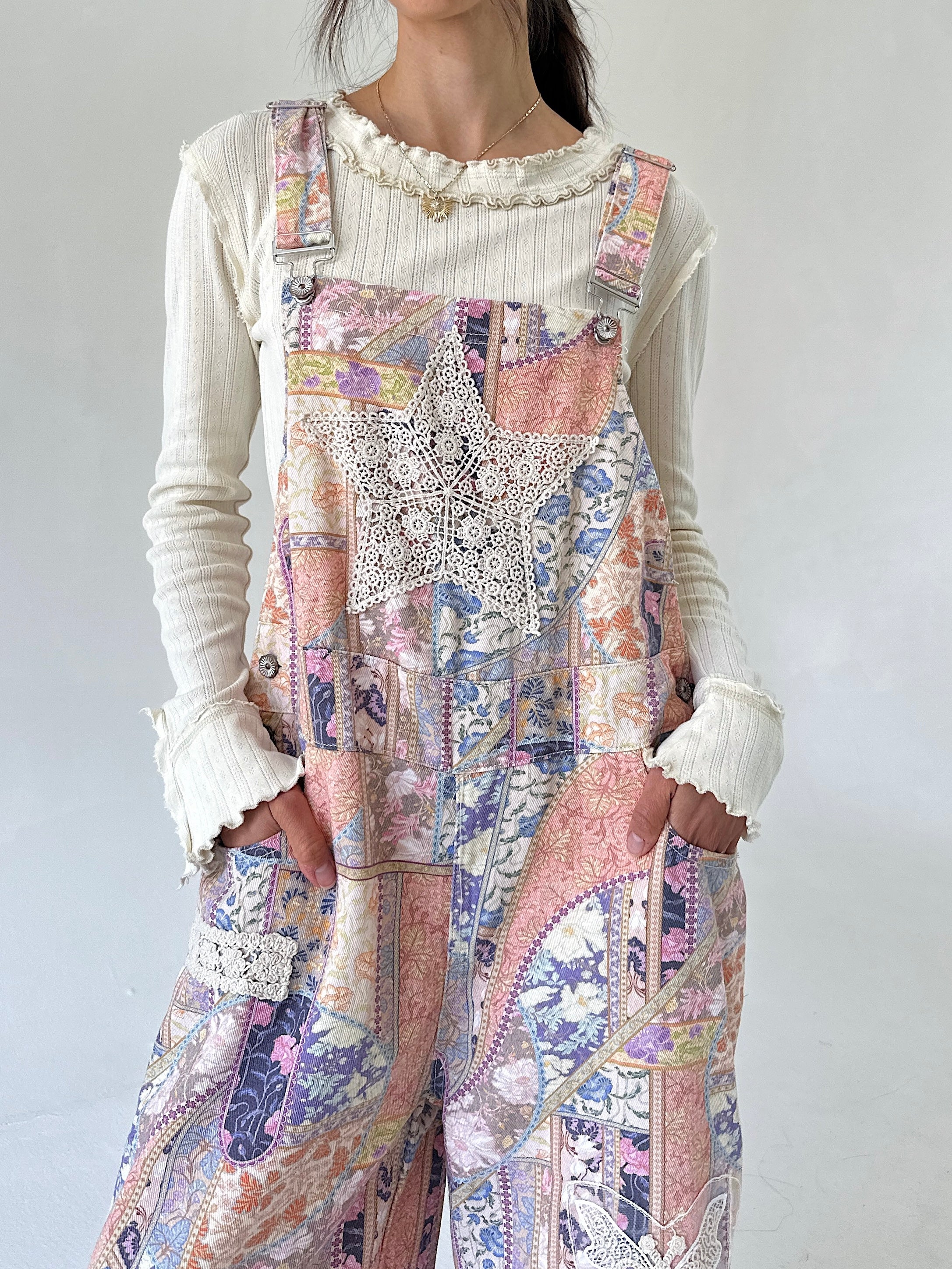 A person wearing a cream ribbed long-sleeve top under pastel patchwork overall pants with lace applique and floral prints in pink, purple, beige, and blue.