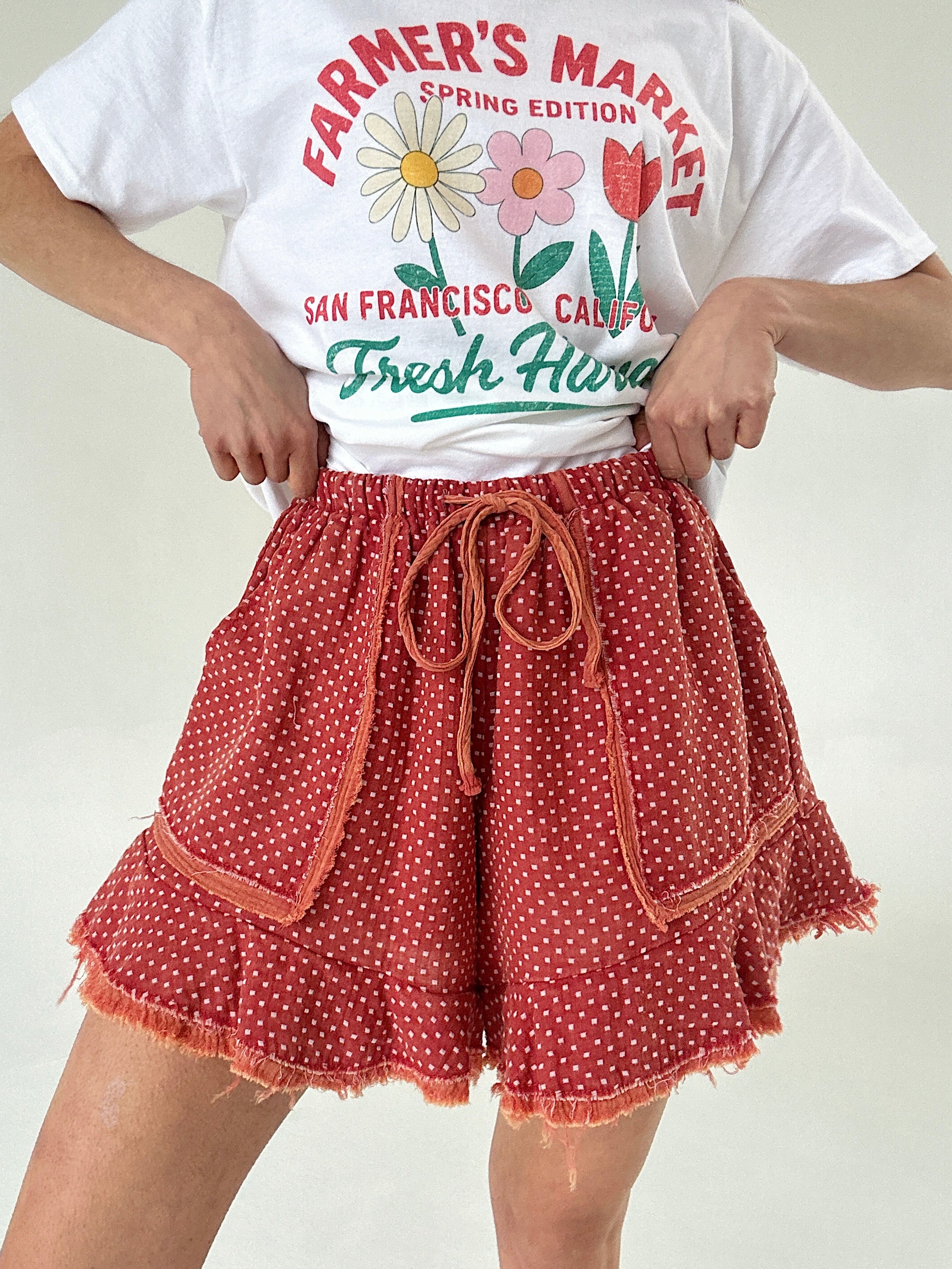 Person wearing a white t-shirt with floral graphics and text, paired with red polka dot shorts on a plain background.
