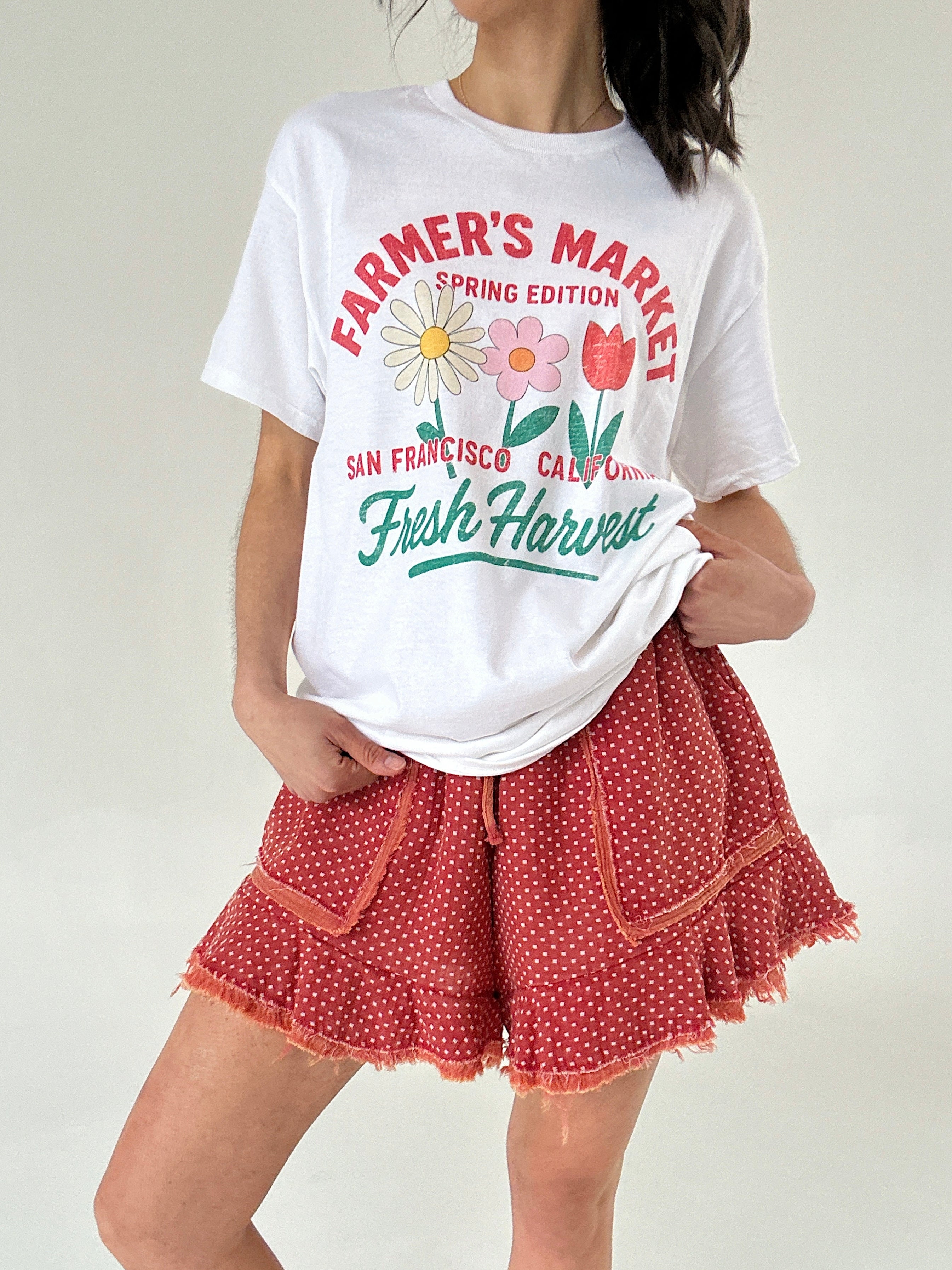 Person wearing a white t-shirt with 'Farmer's Market' text and graphics, paired with red polka dot shorts.