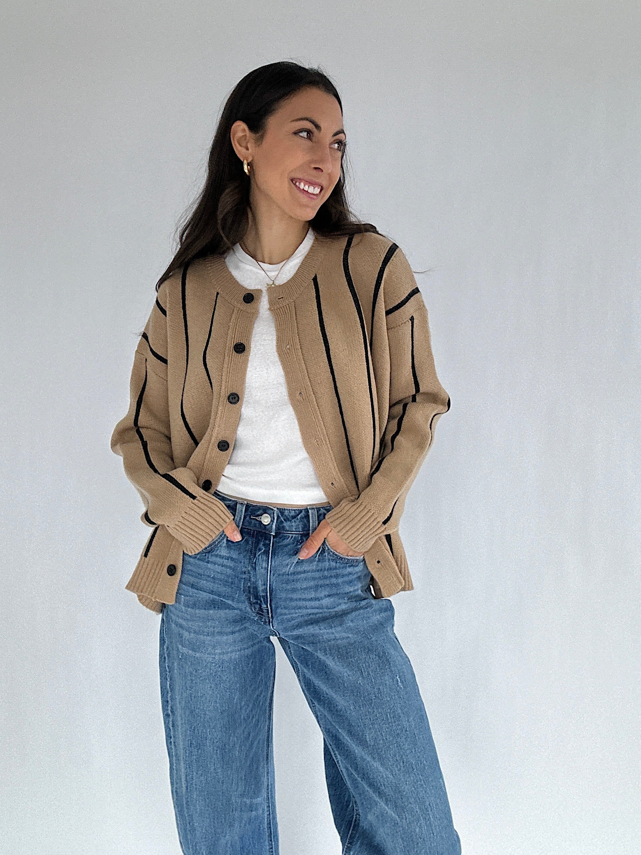 Woman wearing a beige striped cardigan with black buttons, white shirt, and blue jeans against a white background