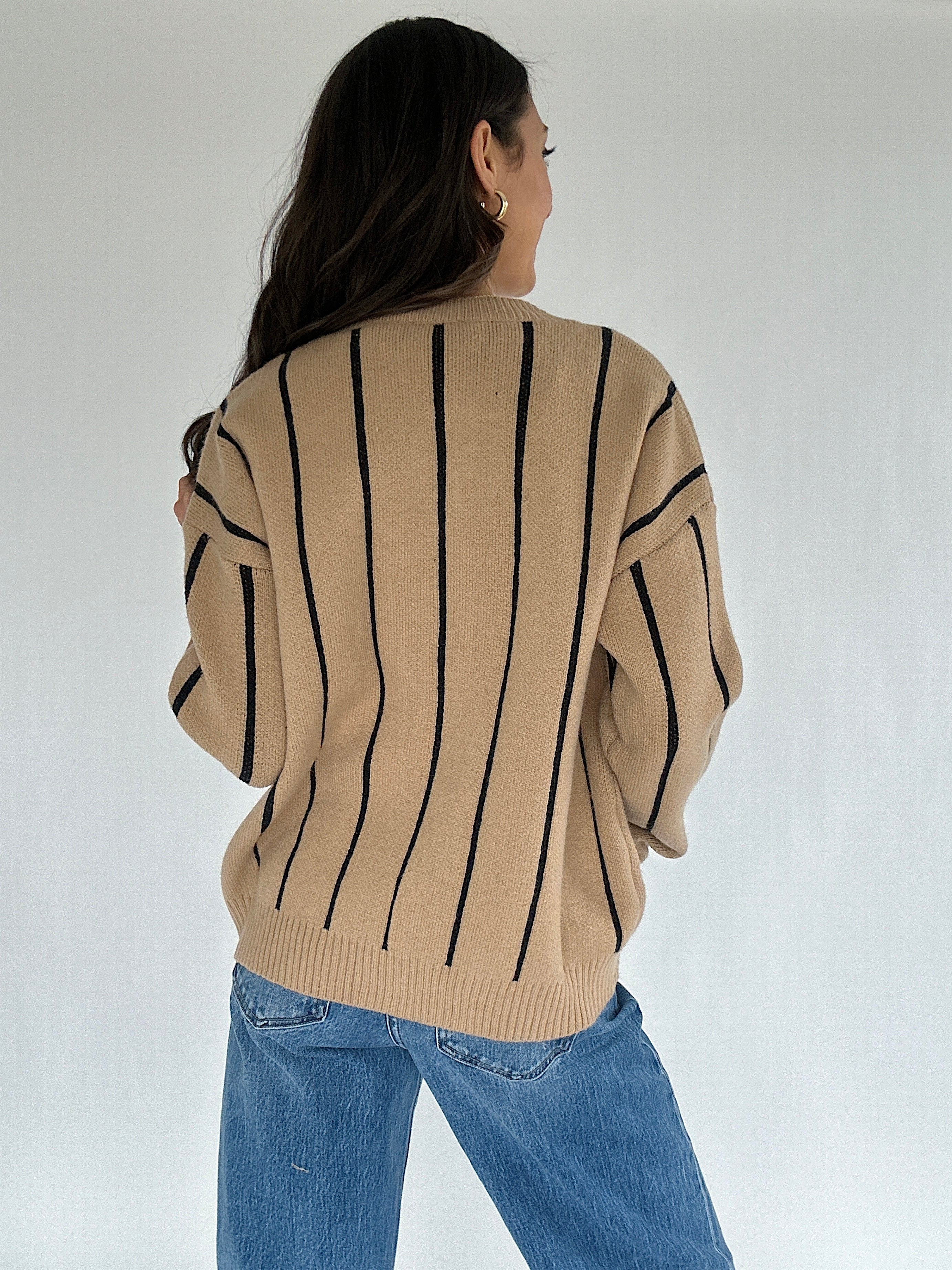 Person wearing a beige sweater with black stripes and blue jeans on a white background showing the back