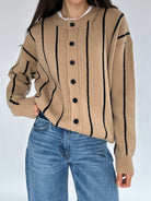 Person wearing a beige sweater with black stripes and buttons, paired with blue jeans on a white background