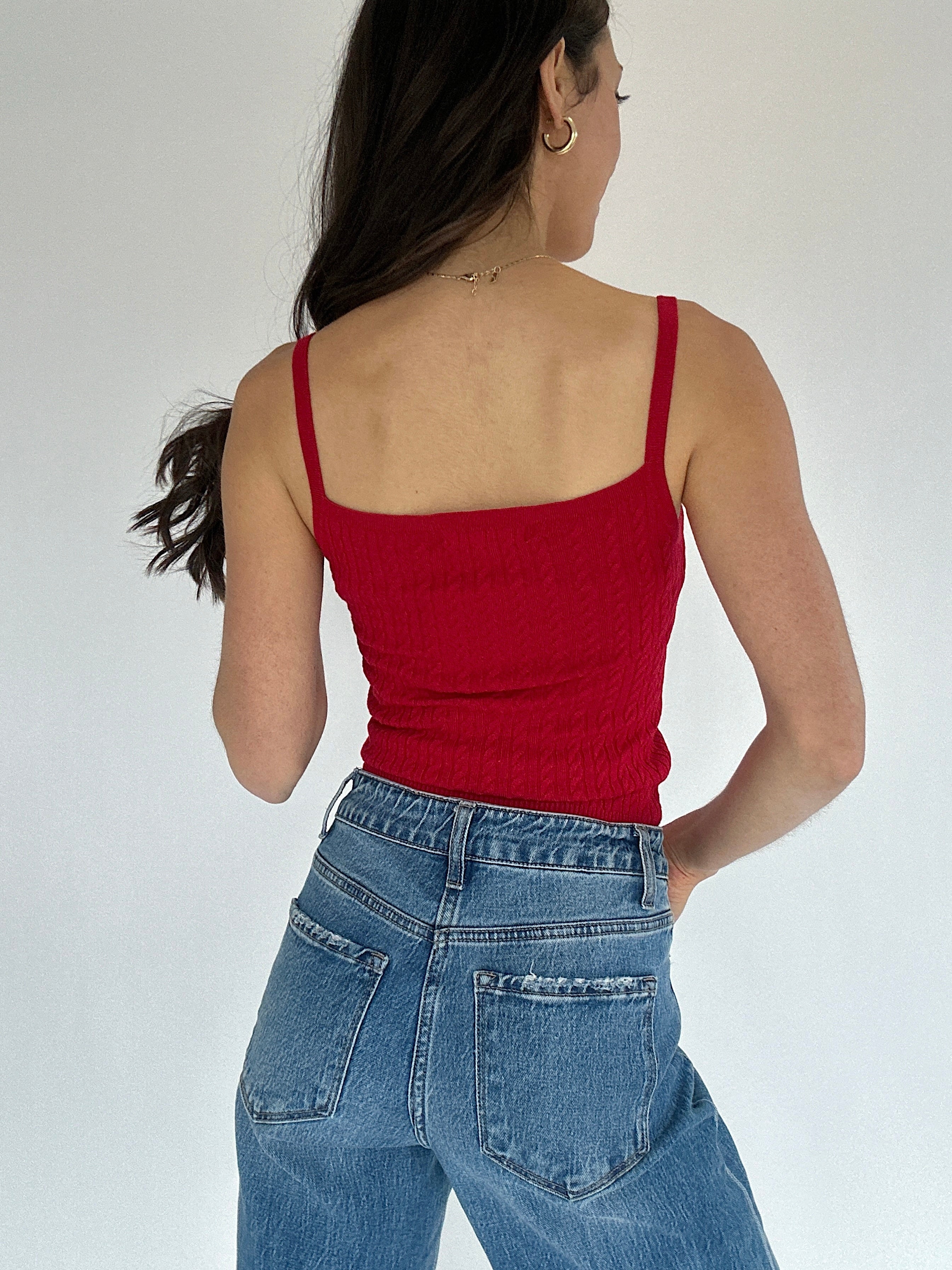 Person wearing a red cable knit tank top and blue jeans against a plain background showing back