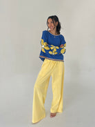 Woman wearing a blue sweater with yellow floral patterns and yellow pants on a plain background