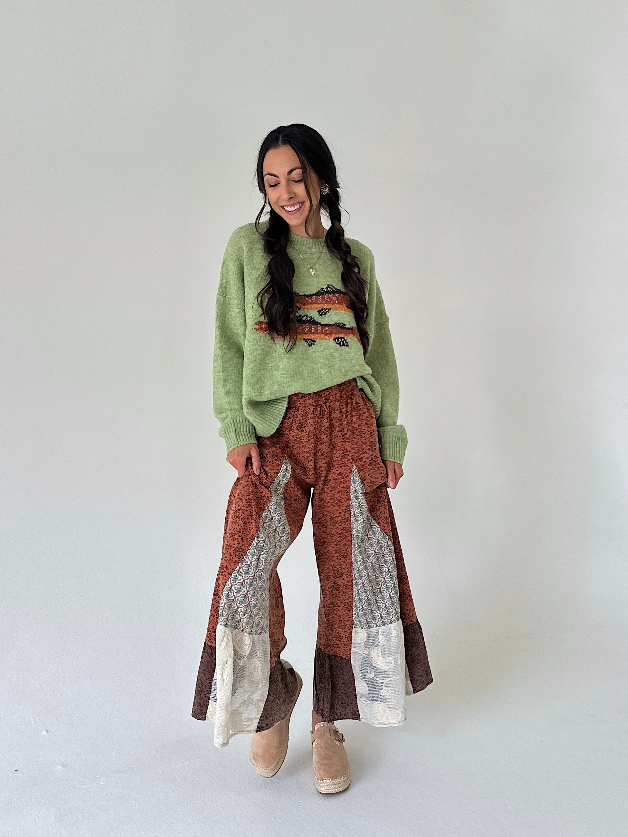 A woman modeling a light green knit sweater with a trout graphic print on the front, paired with brown and beige patterned wide-leg pants and tan clogs, against a plain light gray studio background.