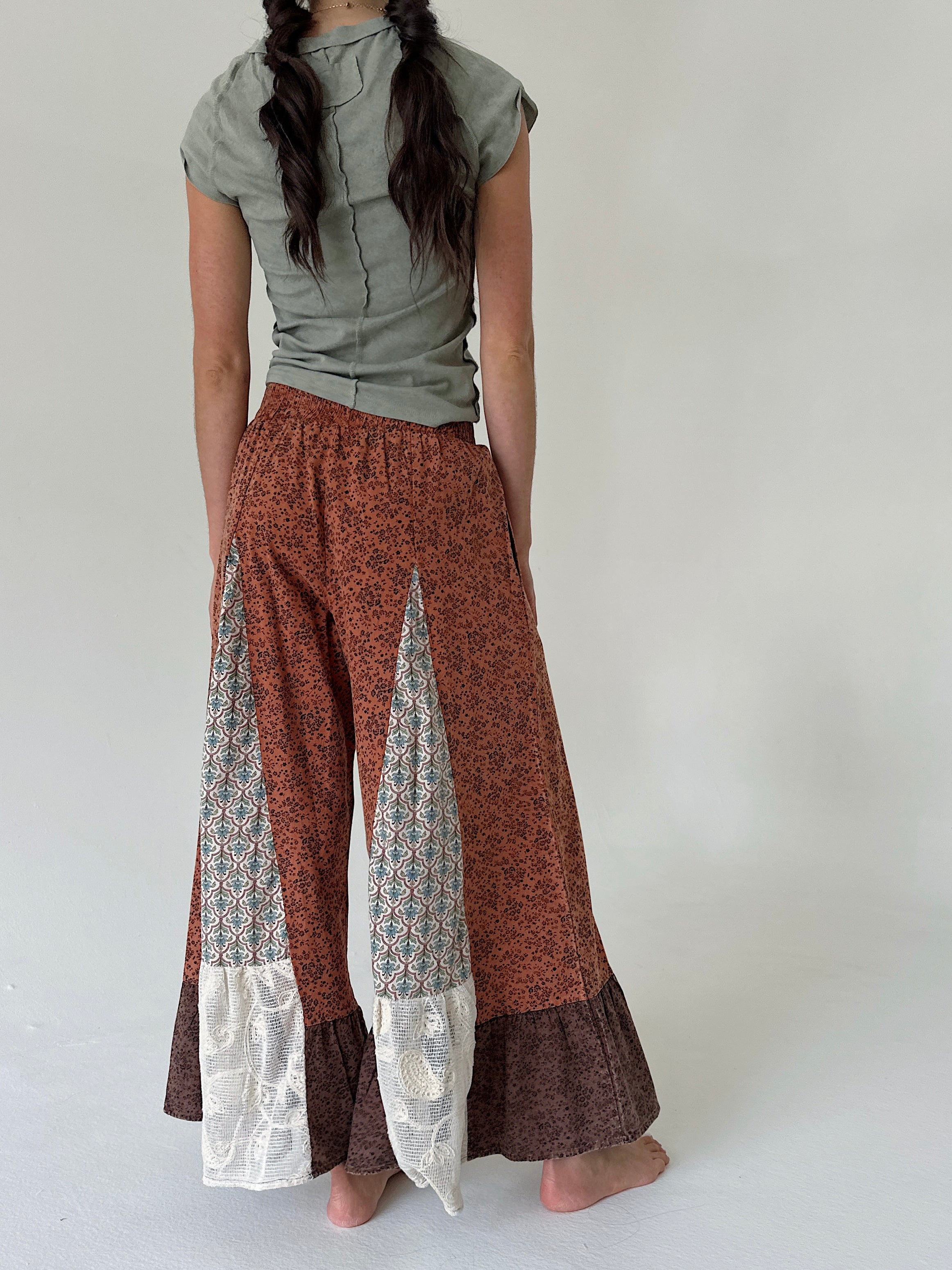 A woman shown from behind wearing wide-leg patchwork pants in rust orange, brown, and white lace, paired with an olive green top, against a plain light gray background.