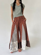 A model wearing wide-leg patchwork pants in rust red, brown, and cream, paired with a sage green t-shirt, standing against a plain off-white studio background.