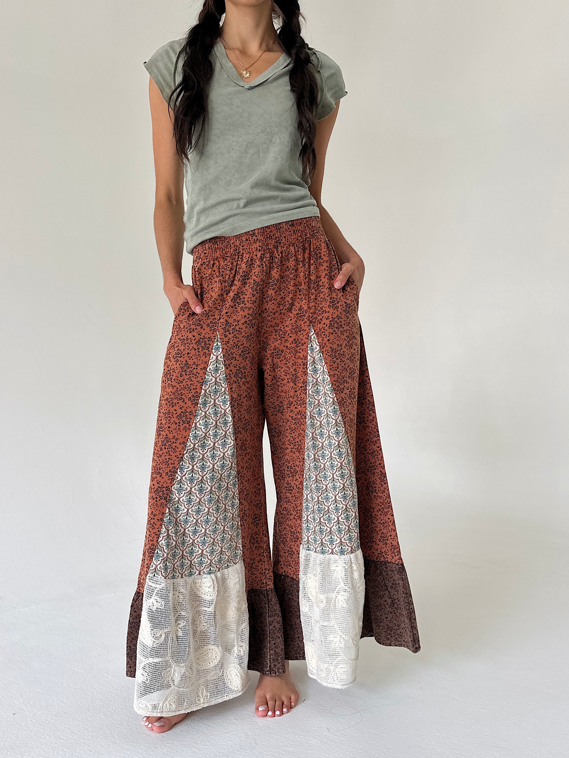A model wearing wide-leg patchwork pants in rust red, brown, and cream, paired with a sage green t-shirt, standing against a plain off-white studio background.