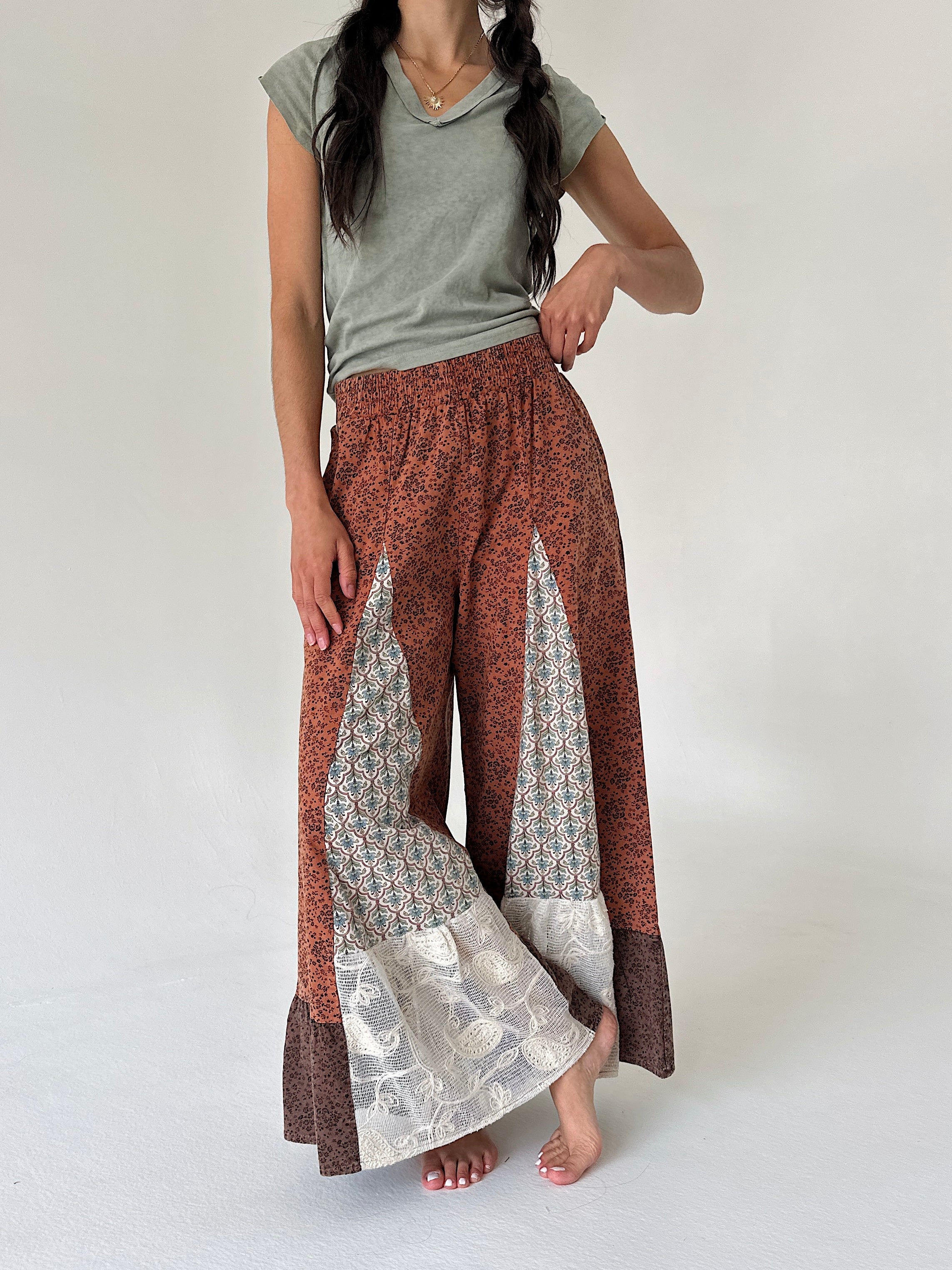 A woman modeling wide-leg patchwork pants with a rust-brown base, geometric patterned panels, lace floral accents, paired with a sage green t-shirt against a plain light gray background.
