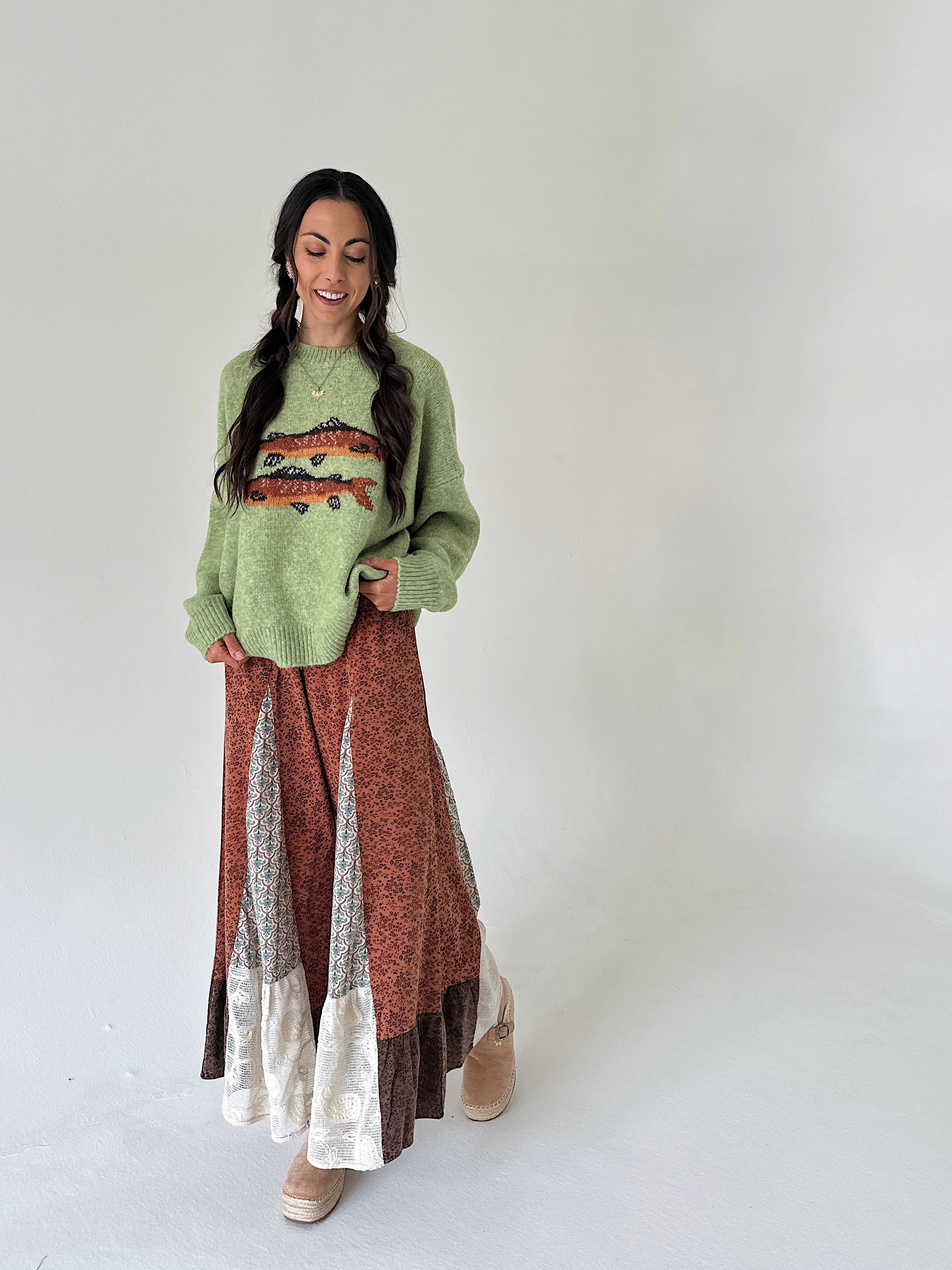 A woman modeling a cozy, long-sleeve knit sweater and a long patchwork maxi skirt. The sweater is a light sage green with a colorful fish design on the chest.