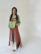 A woman modeling a cozy, long-sleeve knit sweater and a long patchwork maxi skirt. The sweater is a light sage green with a colorful fish design on the chest.