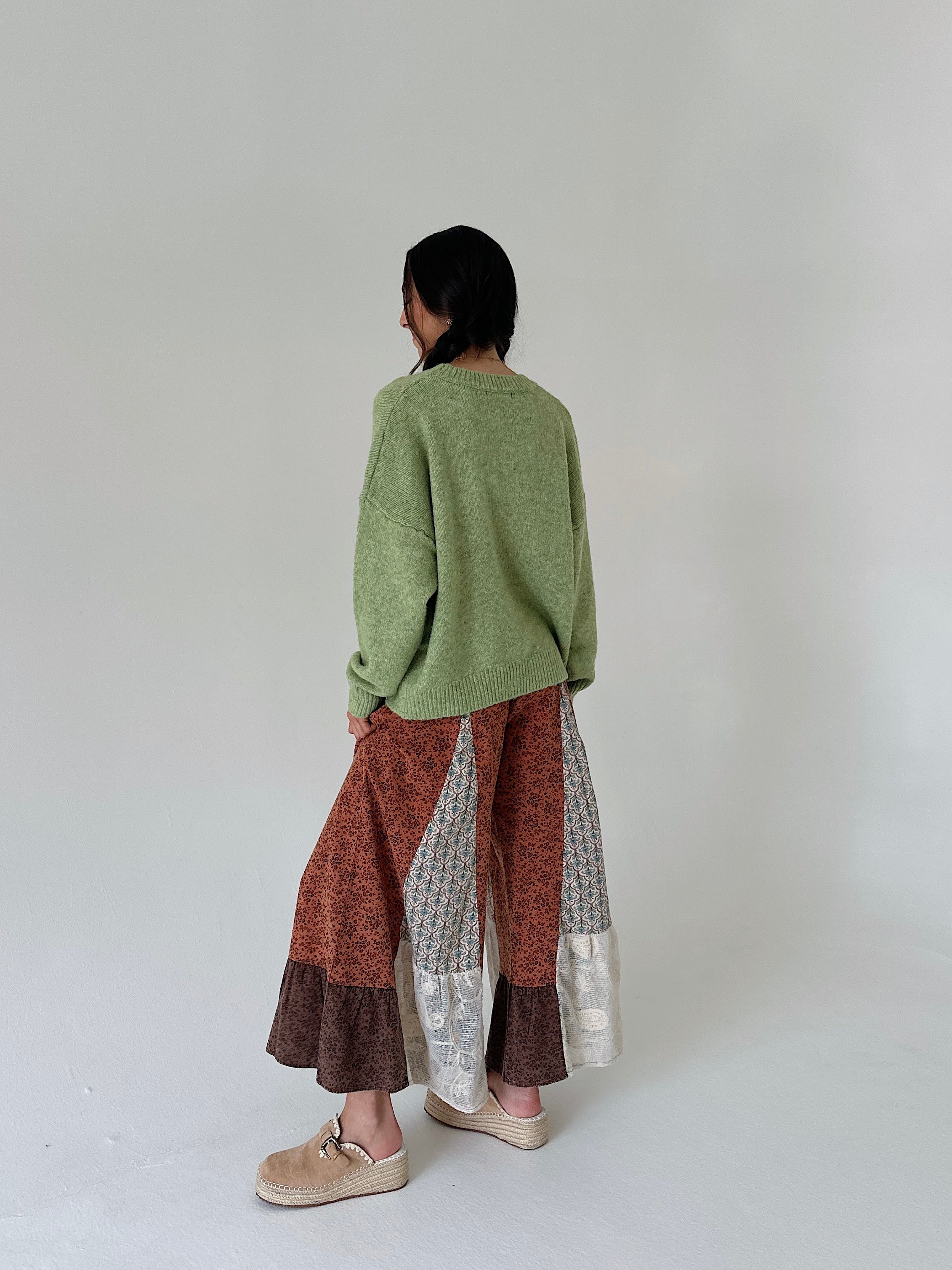 A person shown from the back wearing an oversized sage green knit sweater, wide-leg patchwork pants in rust, brown, and silver-gray, and beige slip-on shoes against a plain light gray studio background.