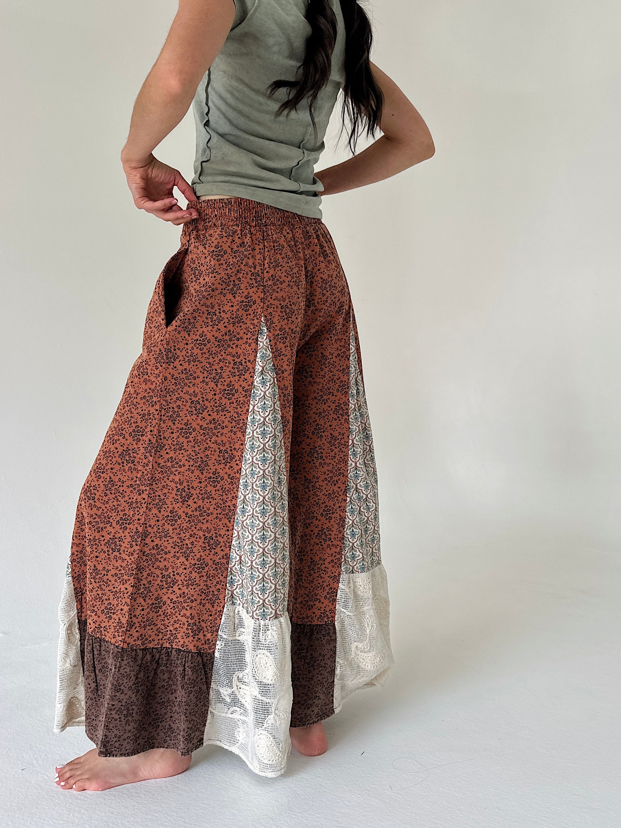 A woman wearing a pair of wide-leg patchwork pants in rust orange, brown, and cream, paired with a sage green top, shown from the back against a plain off-white background.