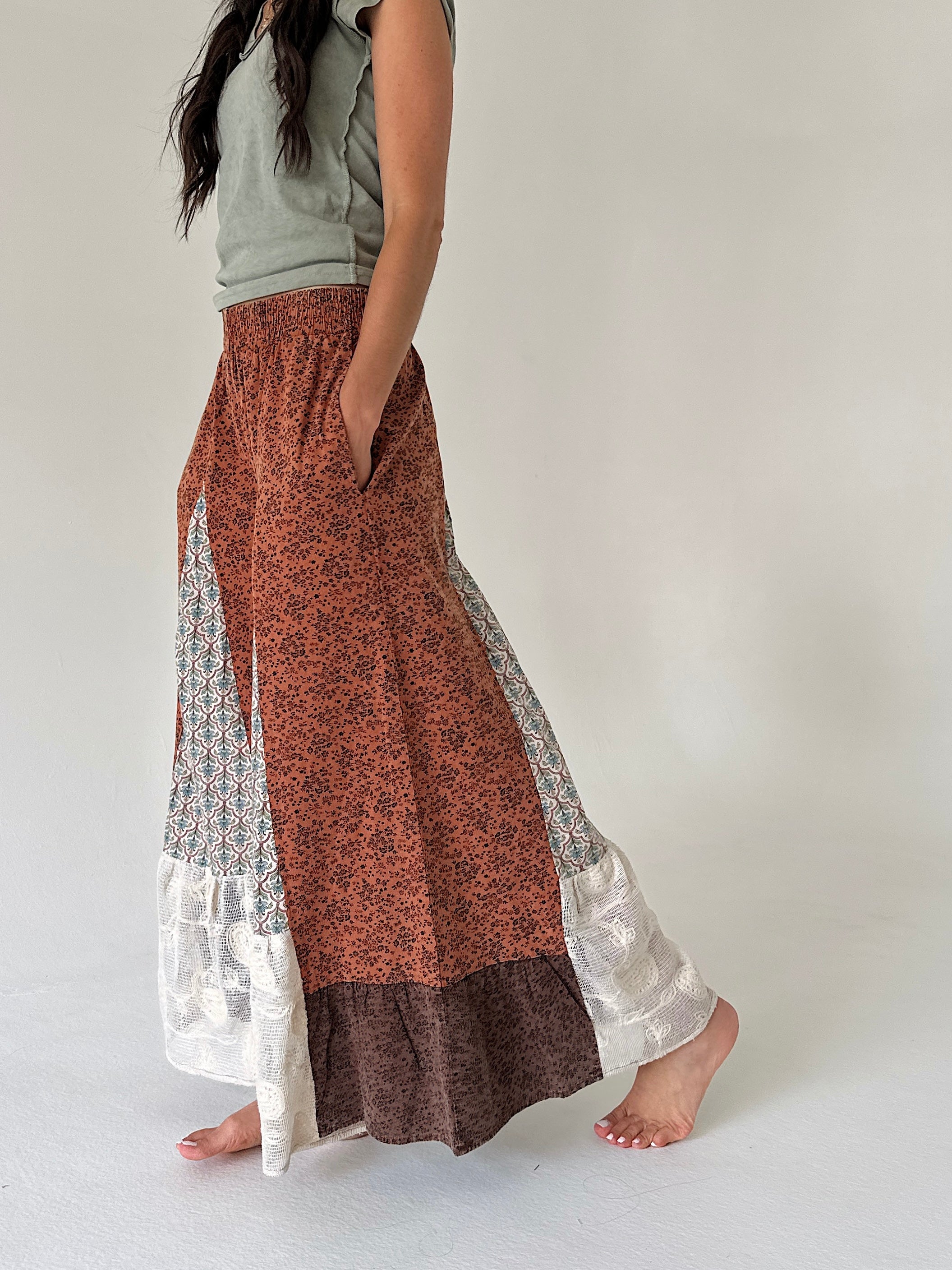 A woman wearing wide leg patchwork pants with rust orange, brown, and white patterns, paired with a light gray top, standing barefoot against a plain off-white background.