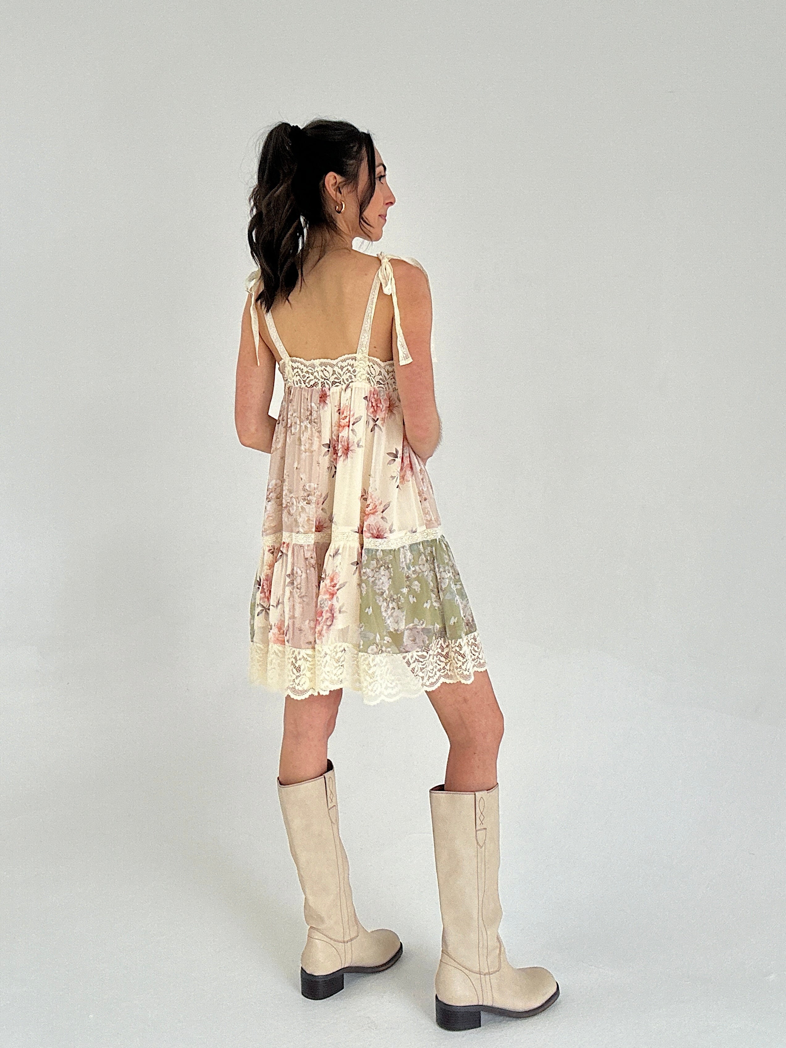Woman wearing a floral dress with lace details and beige boots on a white background showing the back