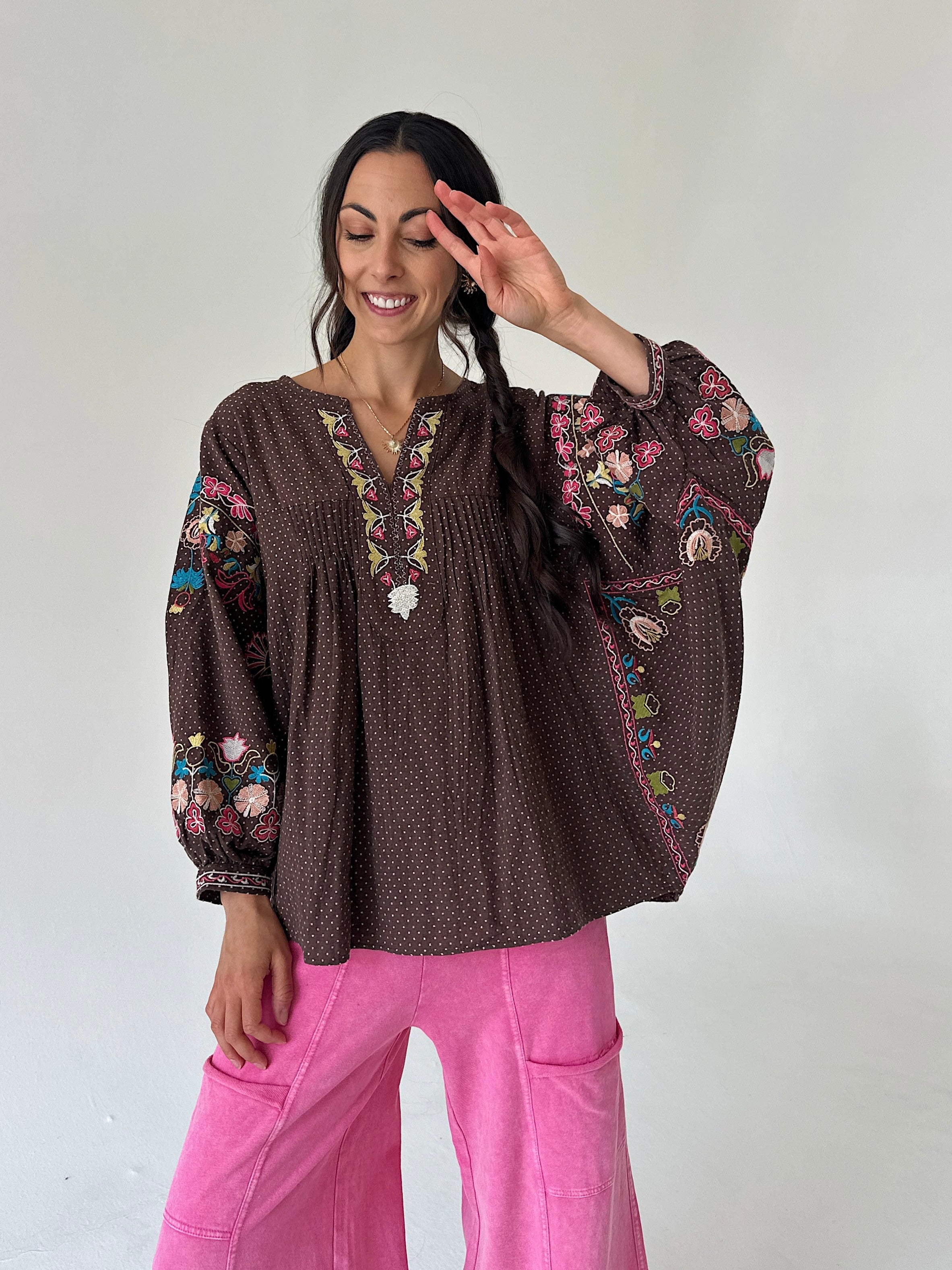A woman wearing a brown bohemian-style embroidered blouse with colorful floral patterns and bright pink pants, posing against a plain light gray wall.